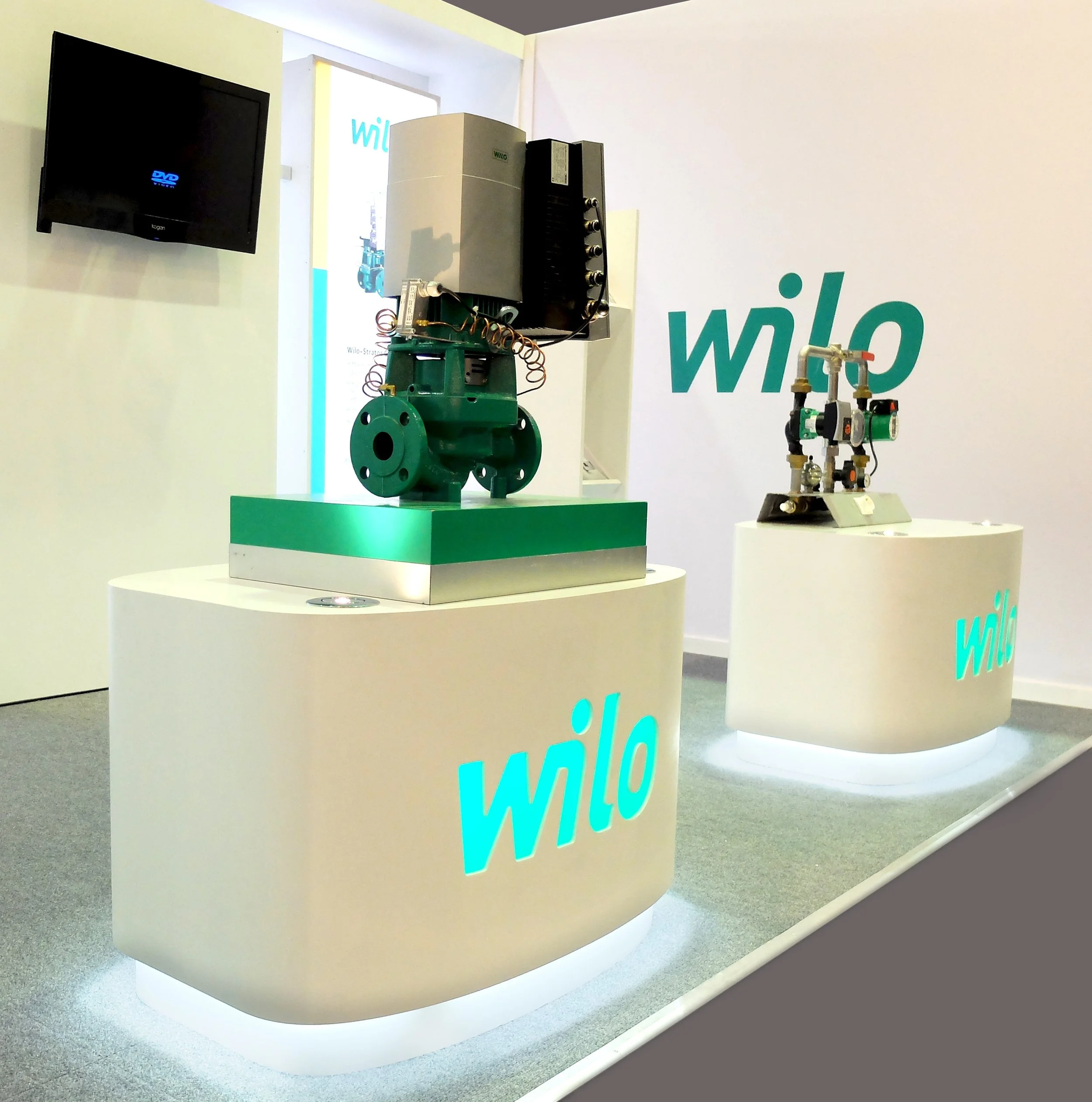 Exhibition display with two industrial components on white pedestals marked with blue and teal 'wilo' logos, featuring a large green valve or pump on the left and a smaller machine on the right, set against a white wall with a black television monitor.