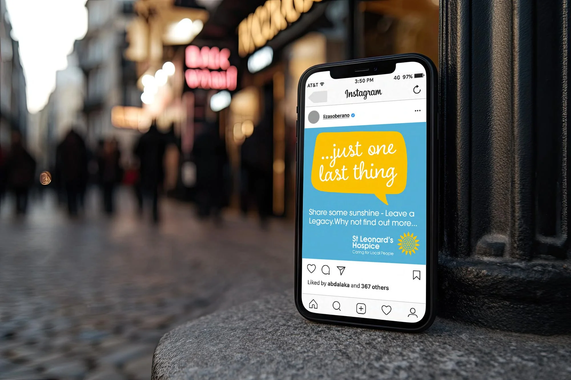 Smartphone displaying a social media post from St. Leonard's Hospice with the message 'just one last thing' in a speech bubble, on a city street corner with blurred pedestrians and storefronts in the background.