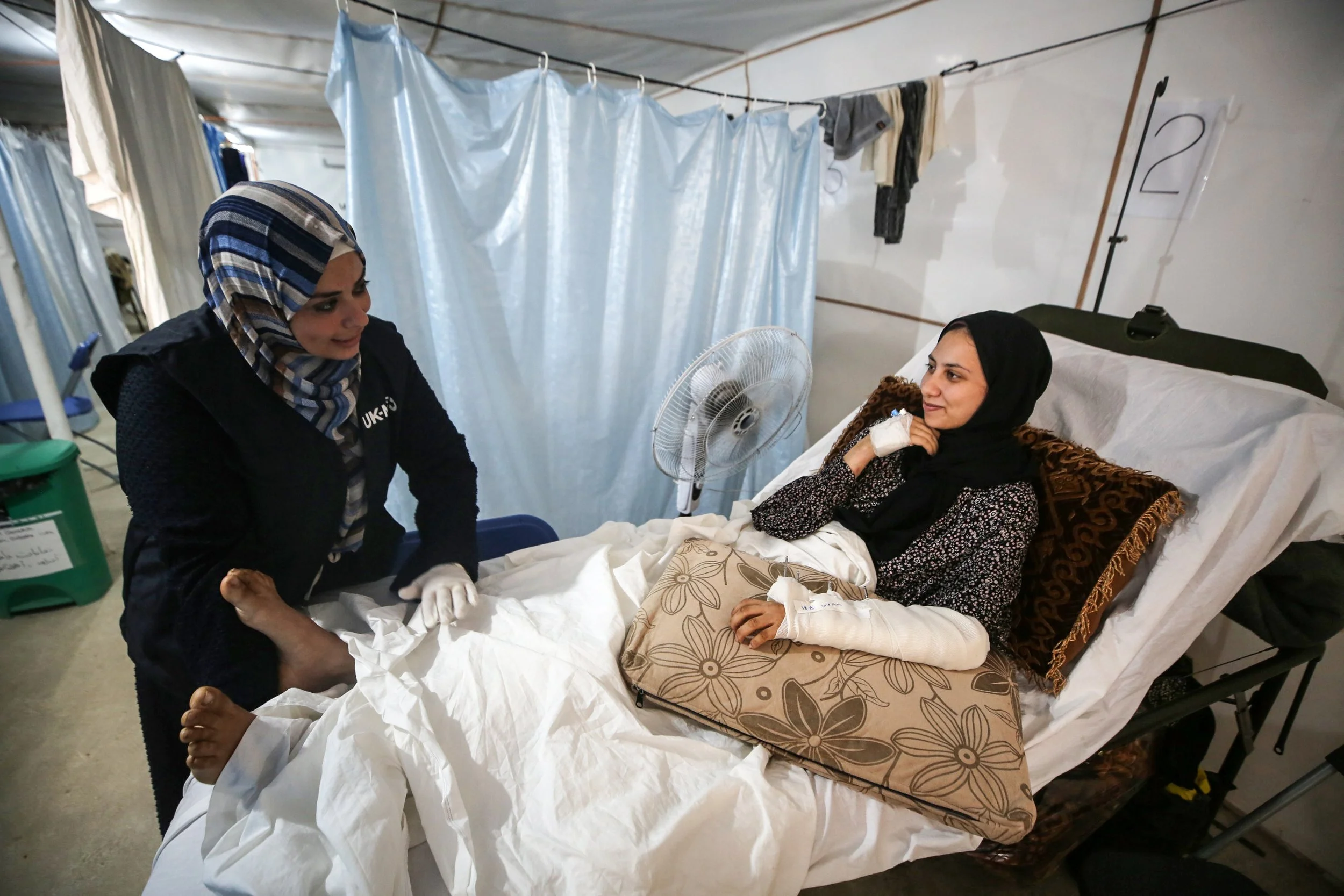 A woman in a hospital bed with a bandaged arm and a head covering, is engaged in conversation with a visitor standing beside her. The visitor also wears a headscarf and is leaning on the bed. The setting appears to be a hospital or medical facility, with curtains and medical equipment visible in the background.