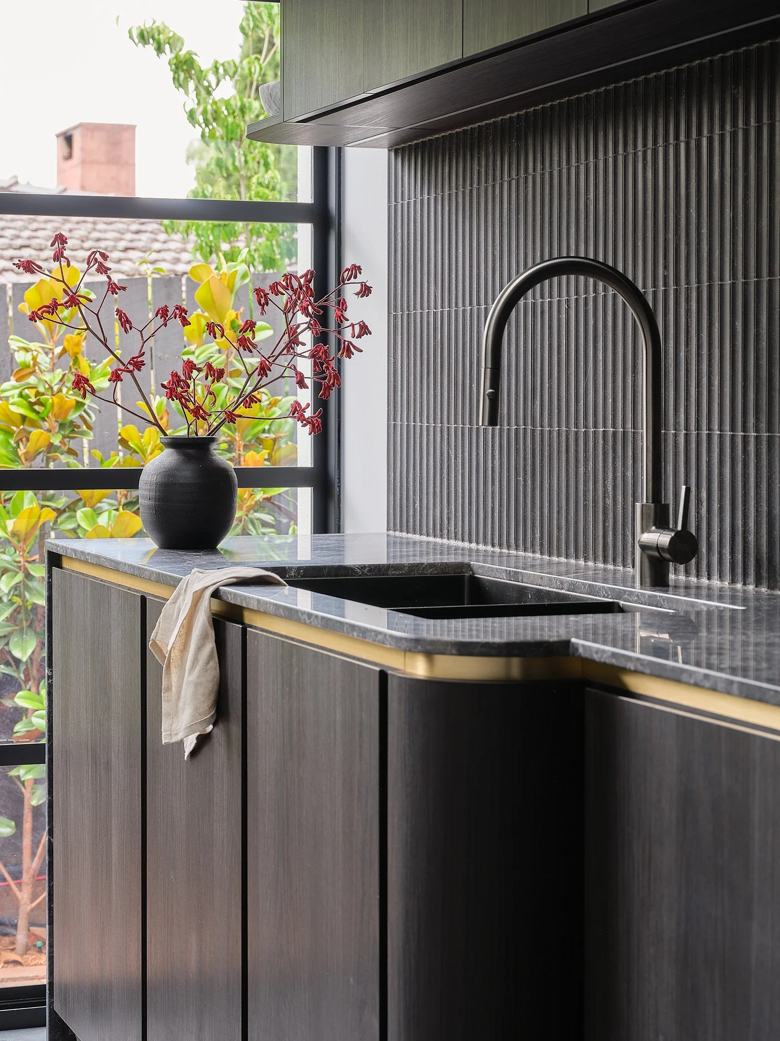 Modern kitchen with black textured backsplash, black faucet, gray natural marble countertop, veneer cabinetry, and a black vase with red and yellow flowers near a large window.