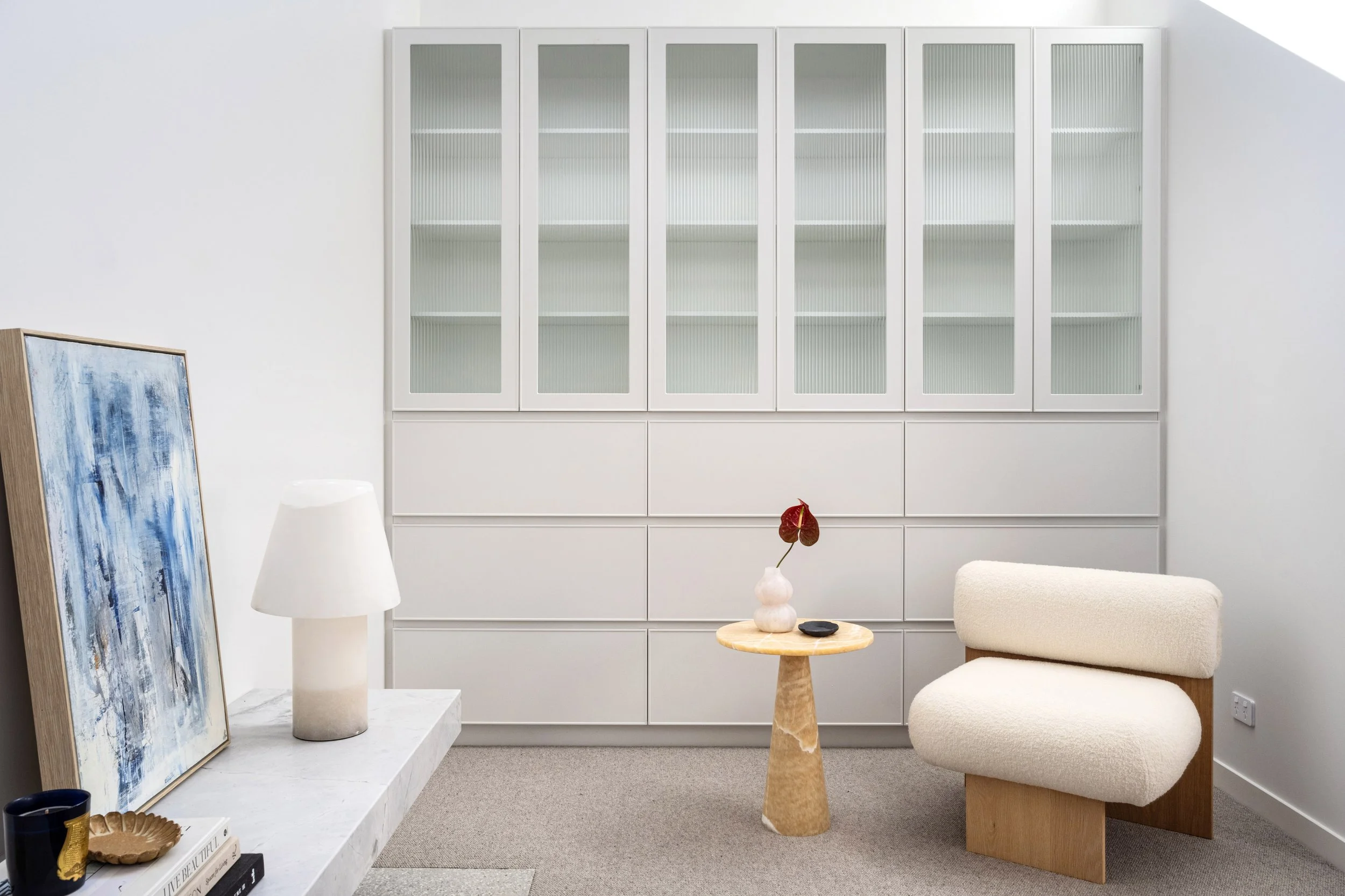 Minimalist living room with white built-in cabinets, a beige armchair, a small wooden side table with a white vase and red leaf, a white marble fireplace with books and decor, a blue abstract painting leaning against the wall, a white lamp, and a gray area rug.