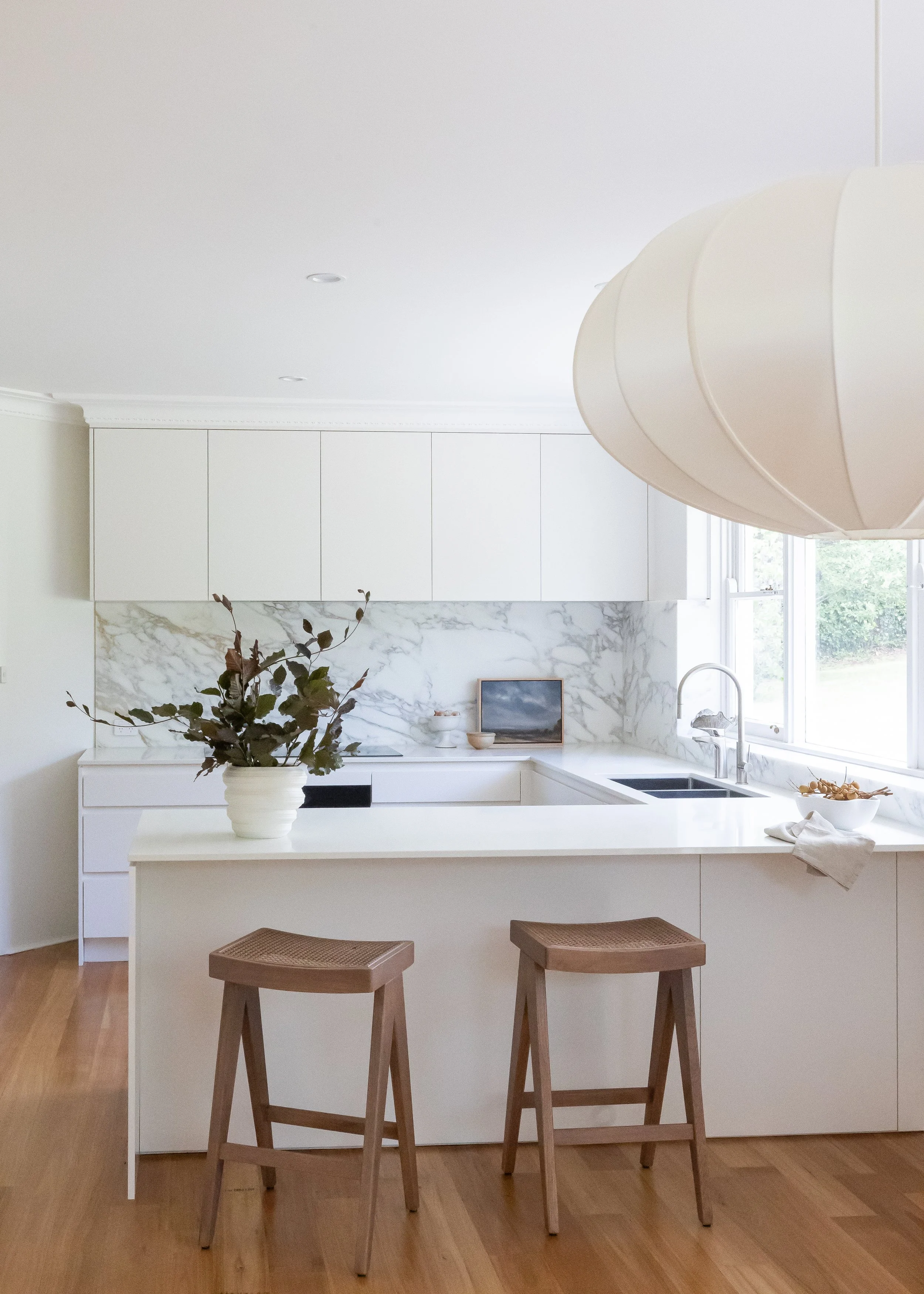 A minimalist kitchen with white cabinetry, a marble backsplash, and a large window. A white kitchen island with a potted plant, stools, and a bowl of fruit. A large, white, hanging lamp is above the island.