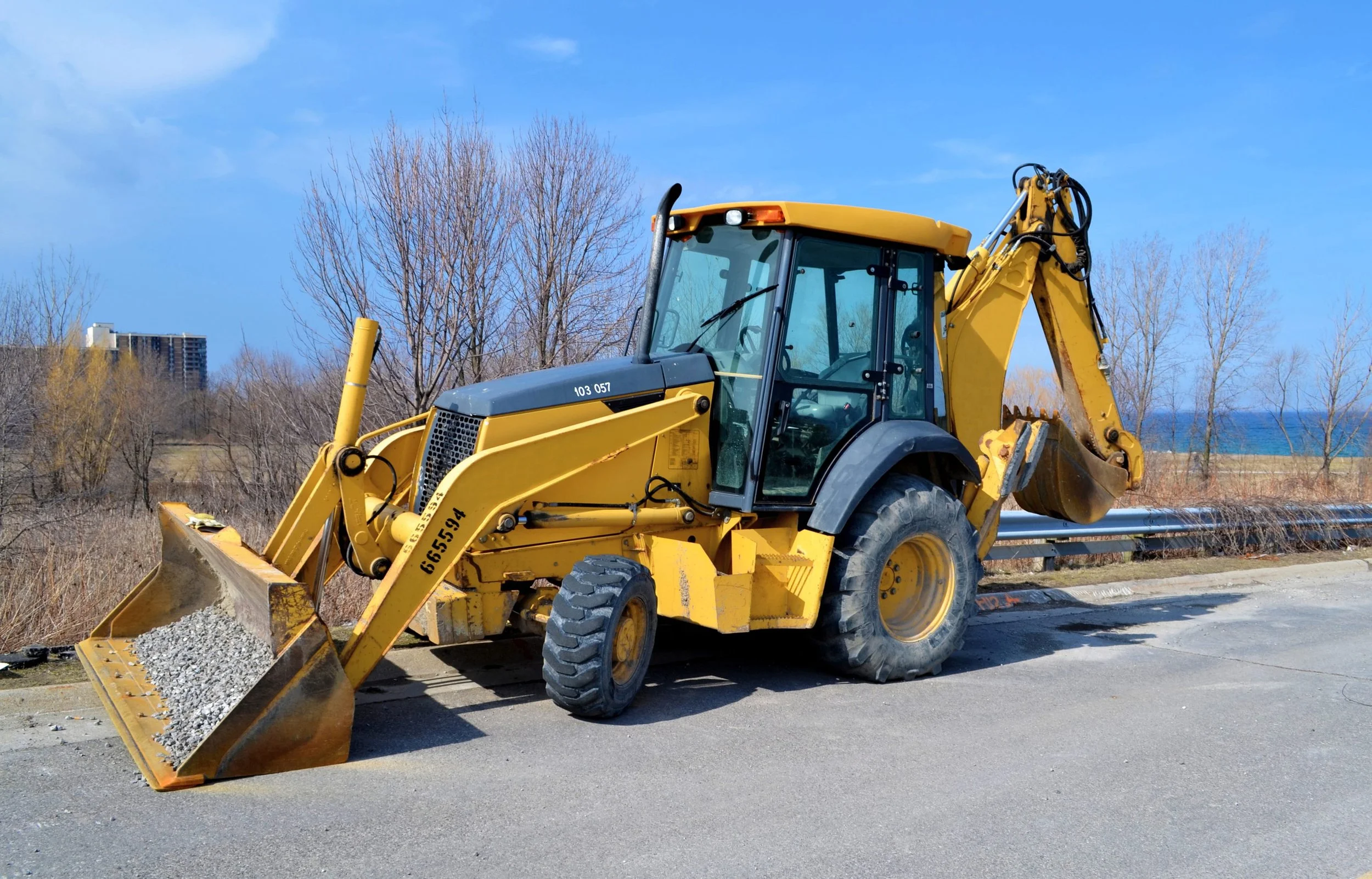 A yellow backhoe loader with a front bucket filled with gravel parked on a road under a blue sky with some clouds. Bare trees and a building are in the background.