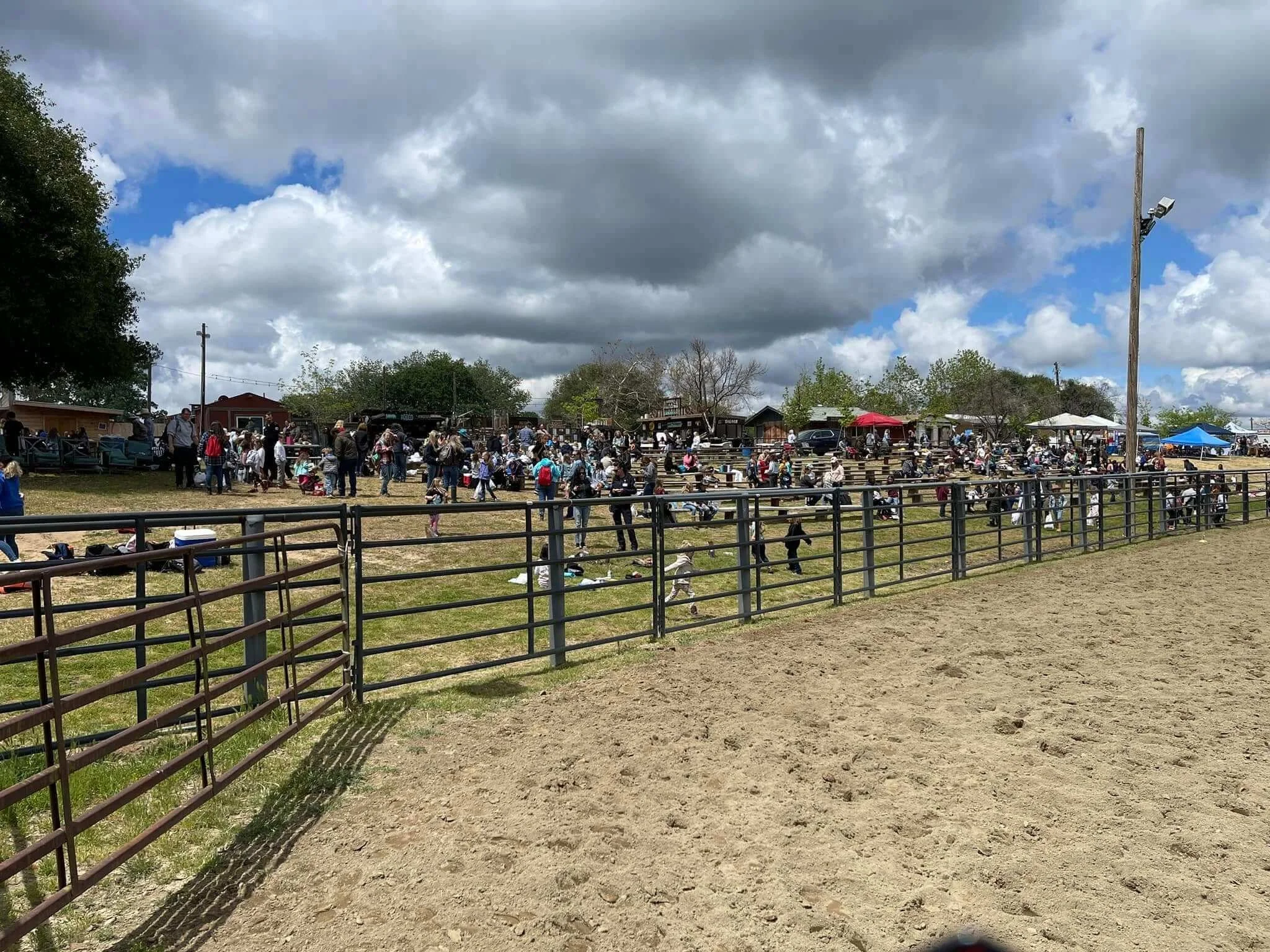 Crowd gathered at a country fair or outdoor event with fencing, tents, and cloudy sky.