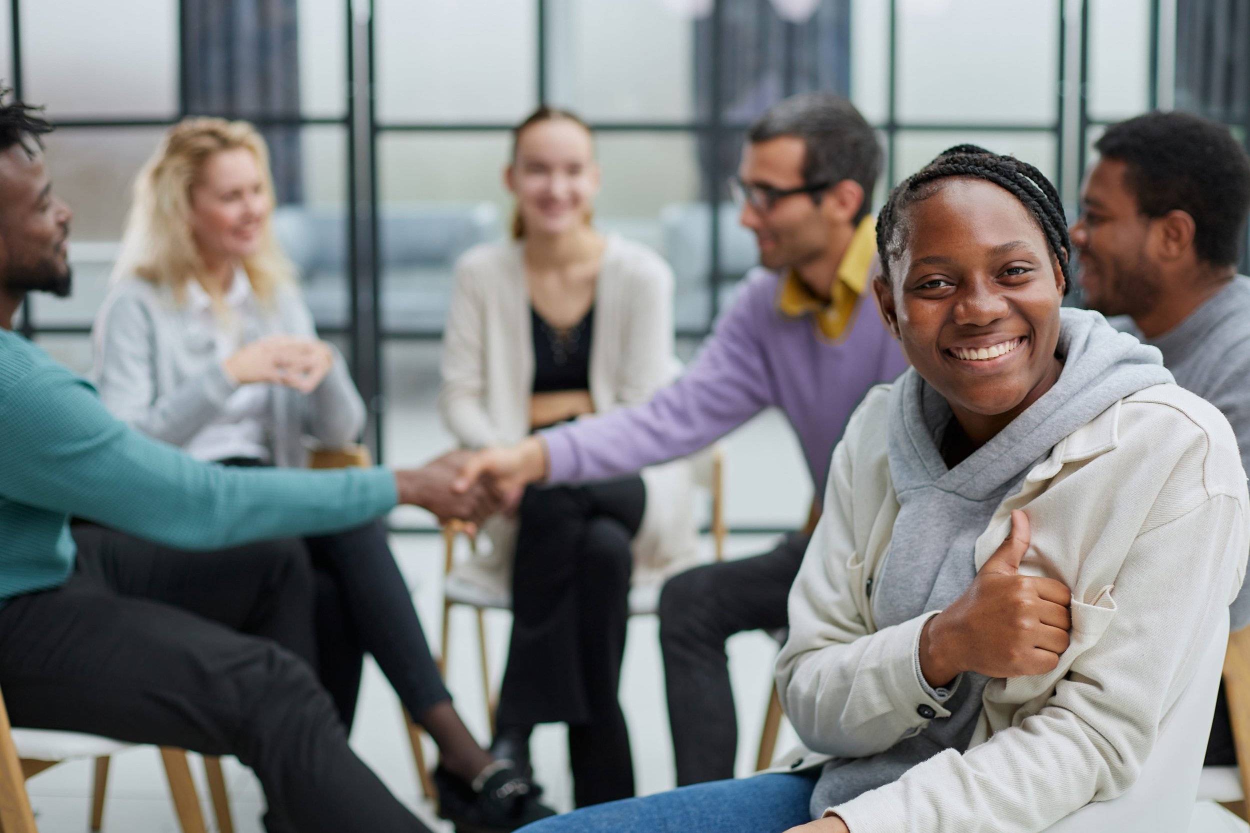 A diverse group of six people in a meeting room, with one woman smiling in the foreground and others shaking hands and talking in the background.