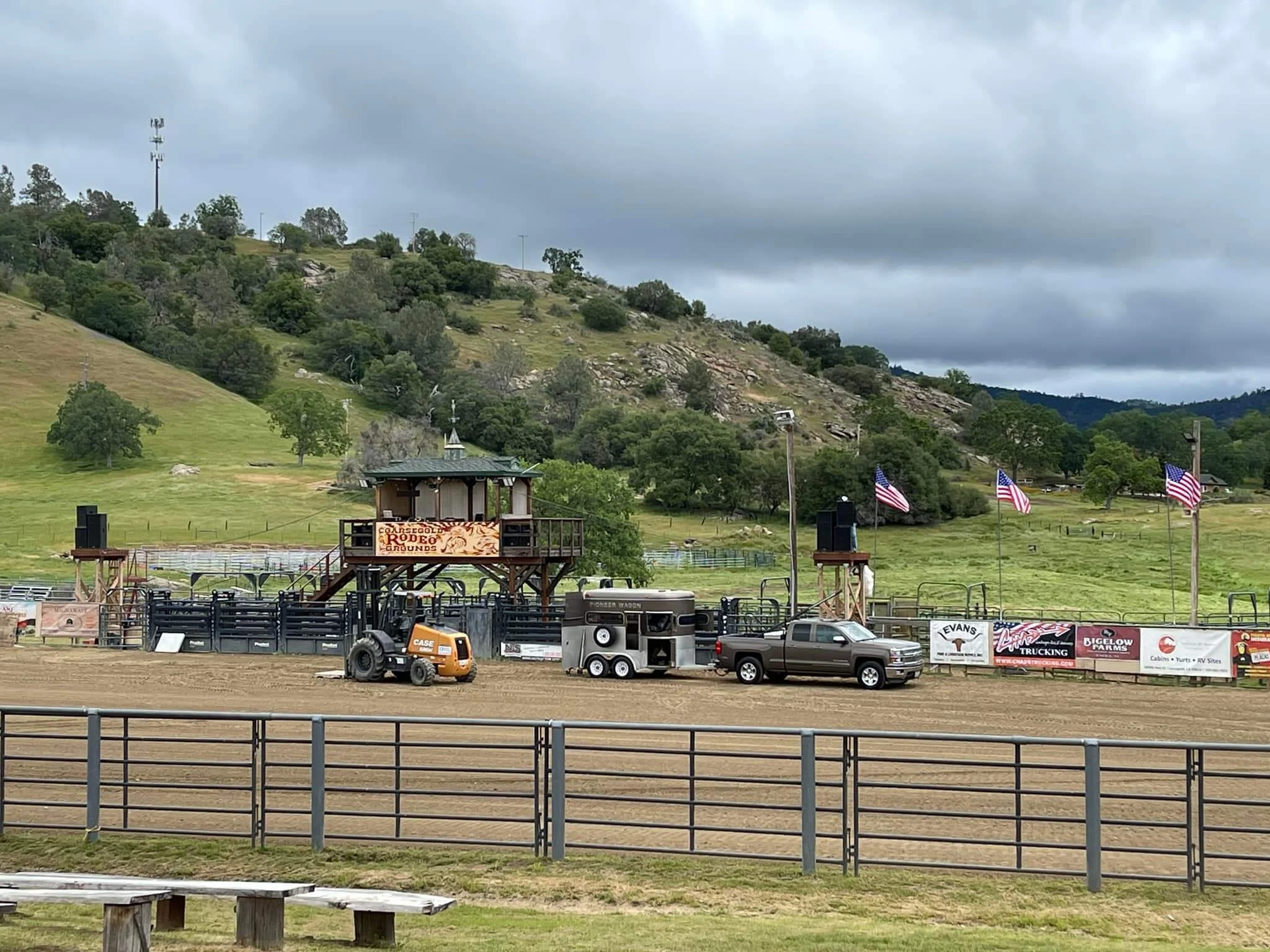 Rodeo arena with dirt track, flags, trucks, and a structure with a sign that reads 'Coles County Rodeo Grounds,' situated in a green, hilly landscape with cloudy sky.