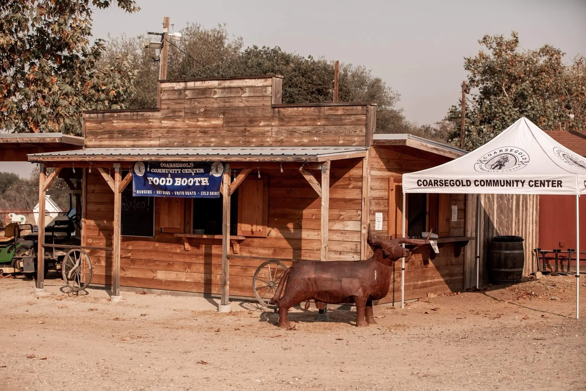 A small wooden building with a sign that reads 'Food Booth' and a white tent with 'Coarsegold Community Center' on it. There is a rusted metal sculpture of a pig with a long snout in front of the building. The setting appears to be outdoors with trees in the background and a dirt ground.