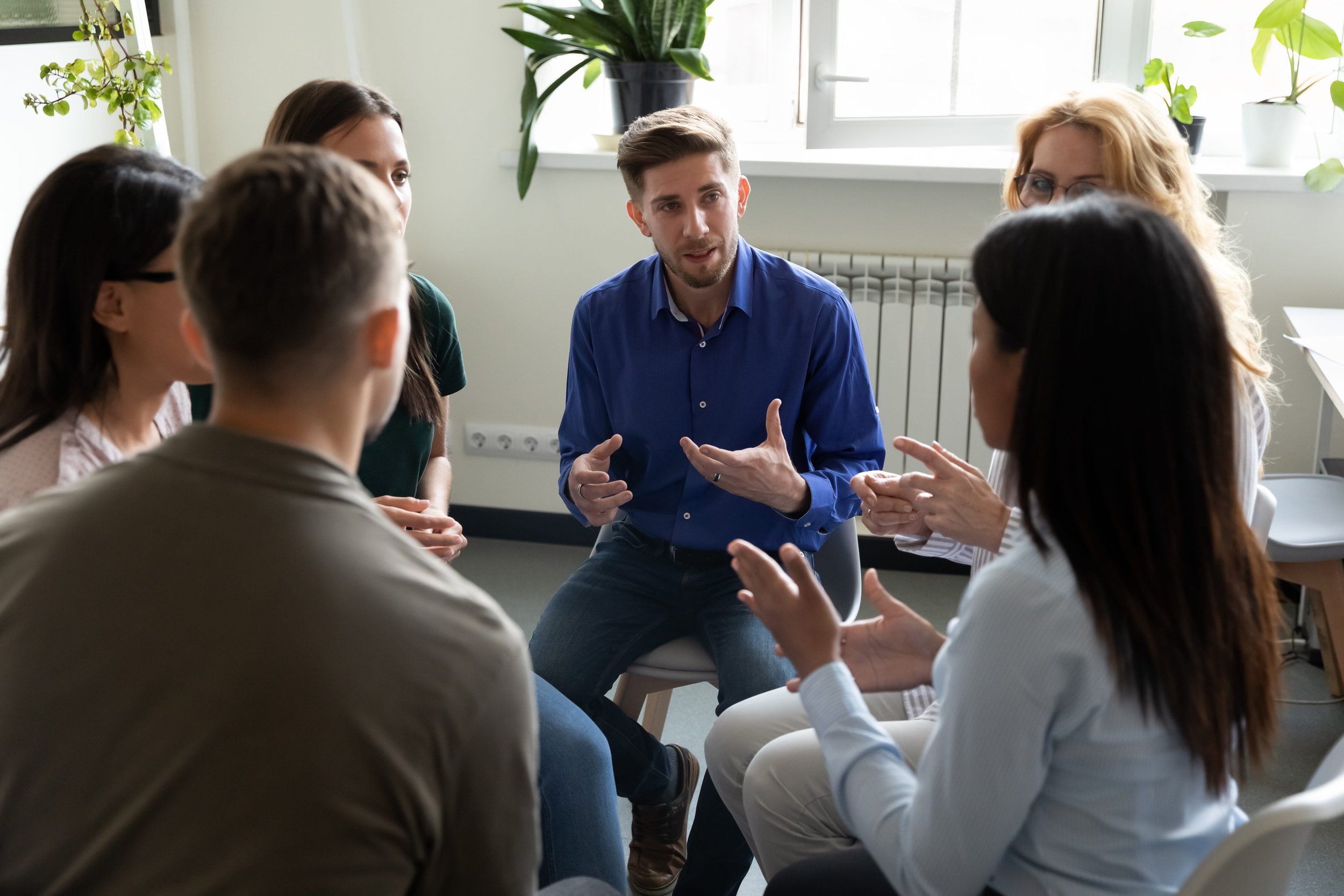 A group of six people engaged in a discussion in an office with natural light and indoor plants.
