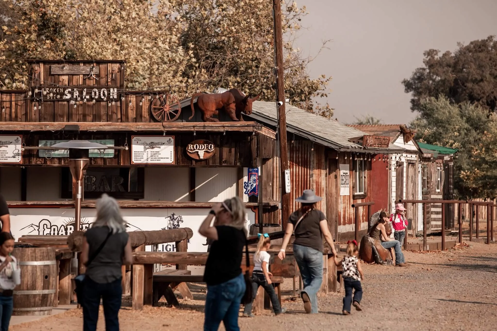 People walking and sitting outside a rustic wooden building with signs, including a donkey statue on the roof, trees in the background, and dusty ground.