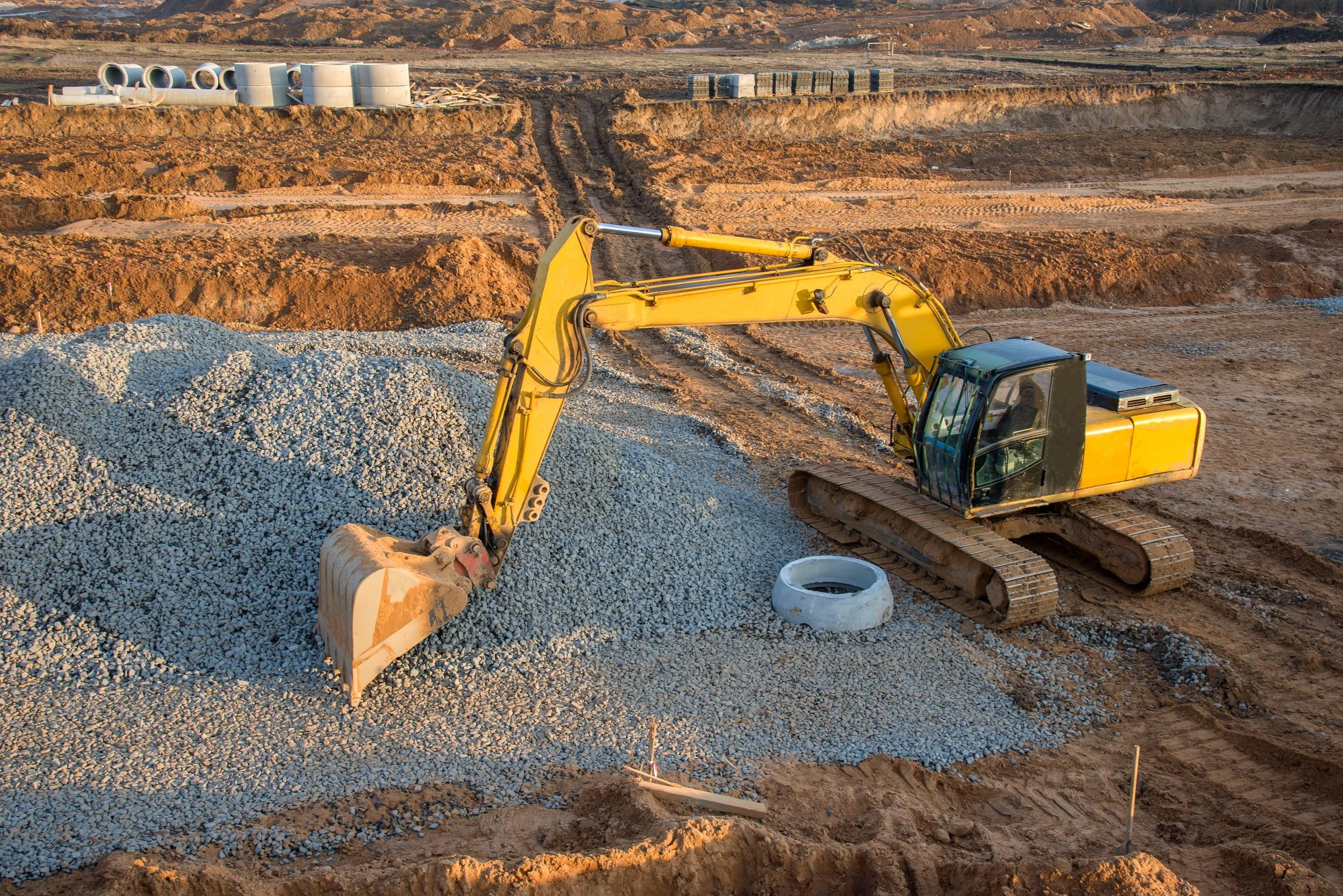 Yellow excavator at a construction site, moving gravel with a large bucket, with dirt and construction materials in the background.