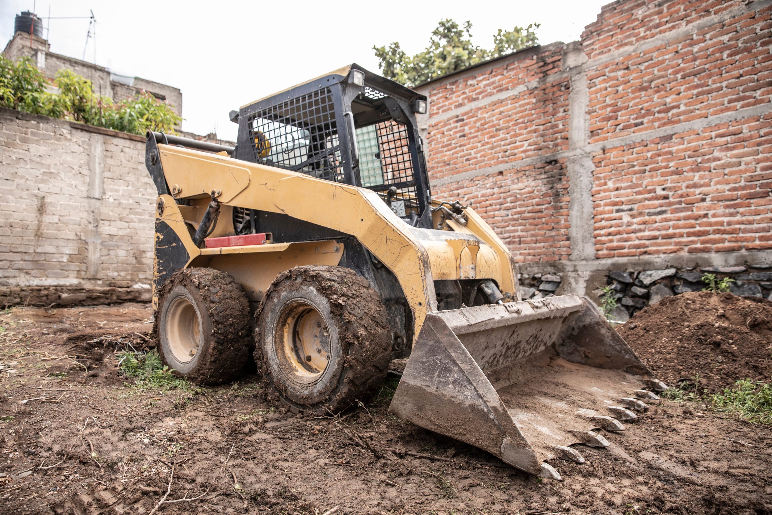 A yellow construction loader with muddy tires on a dirt construction site, with brick walls in the background.