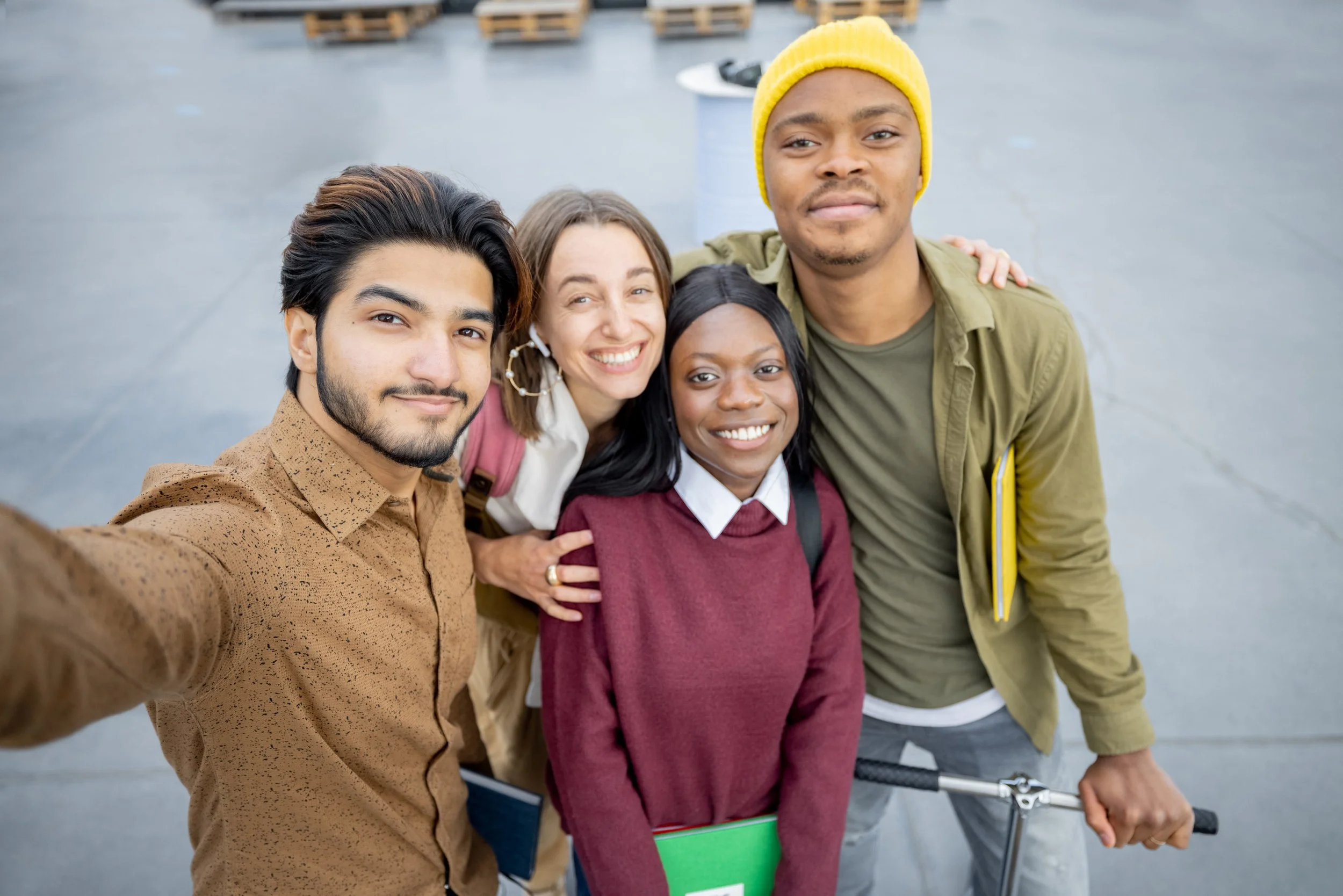 A diverse group of four young friends taking a selfie together outdoors, smiling. One person is holding a scooter, another has a notebook, and they are standing on a paved area.