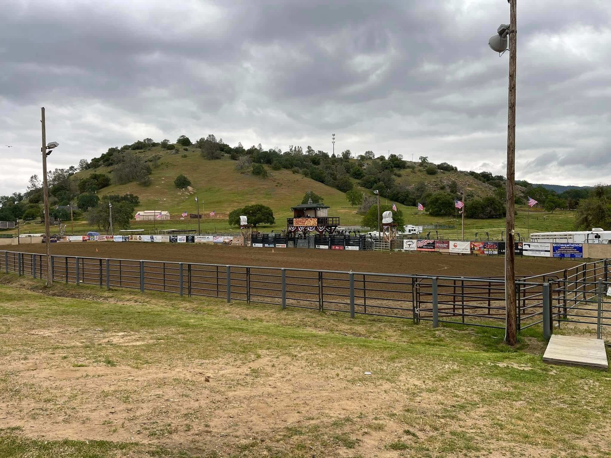 Empty rodeo arena with dirt ground, surrounded by metal fencing, with a small central stage, flags, and hills in the background under cloudy sky.