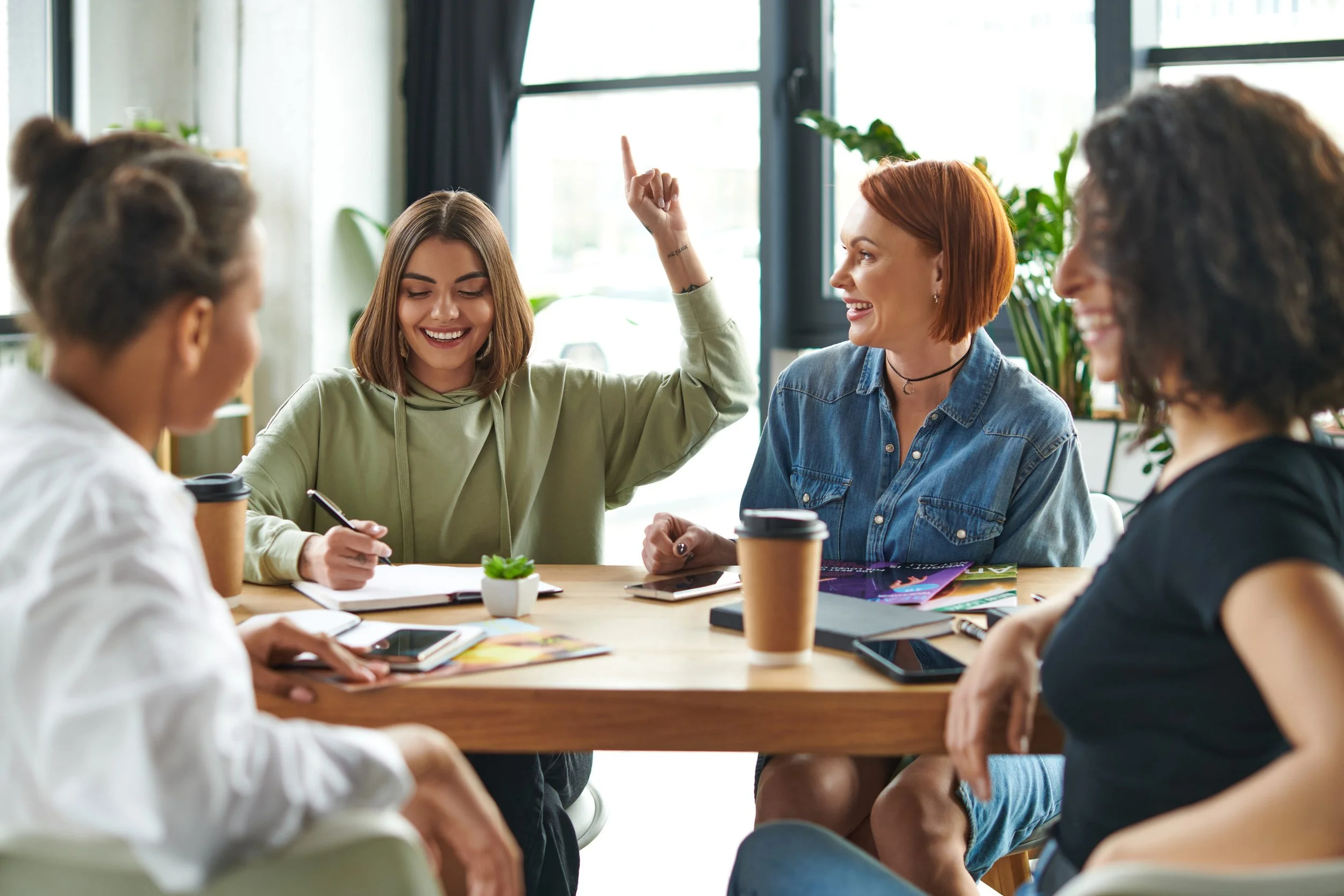 A group of women sitting around a table in a bright, modern office, engaging in a conversation with one woman raising her hand.