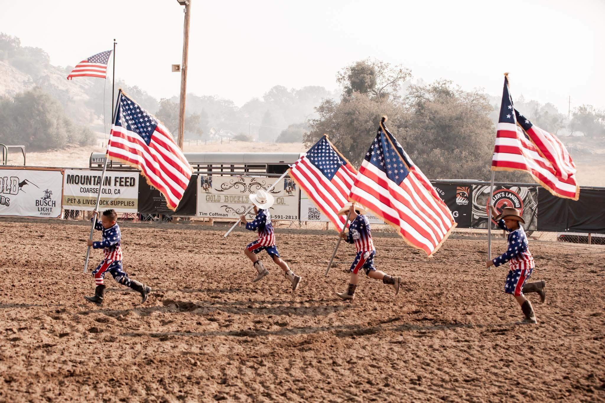 Children dressed in patriotic American flag-themed clothing running on a dirt field while holding large American flags.