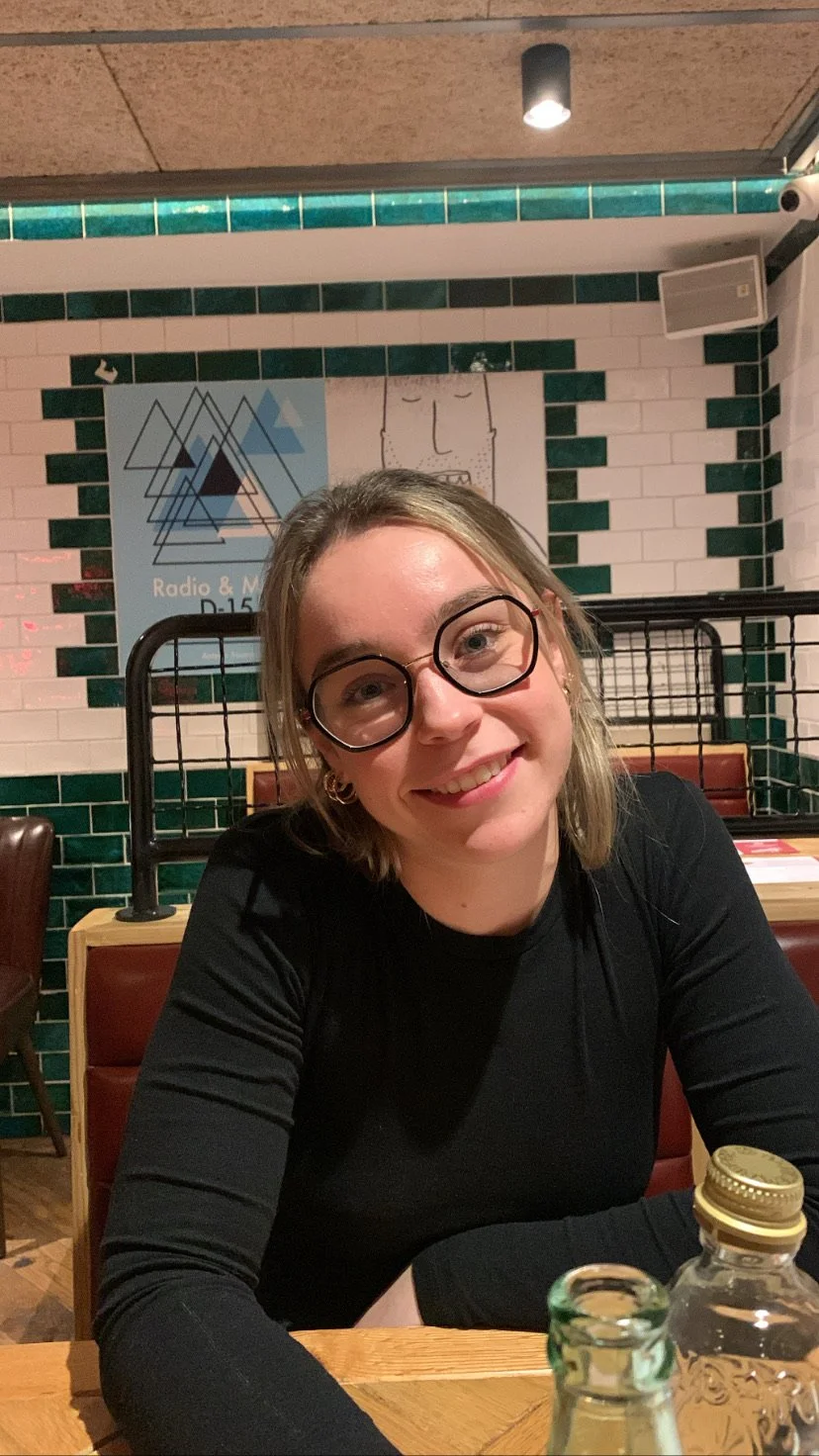 Mujer sonriendo con gafas de marco grande, sentada en una mesa en un restaurante, con mosaicos verdes y blancos en la pared y un cartel en el fondo.