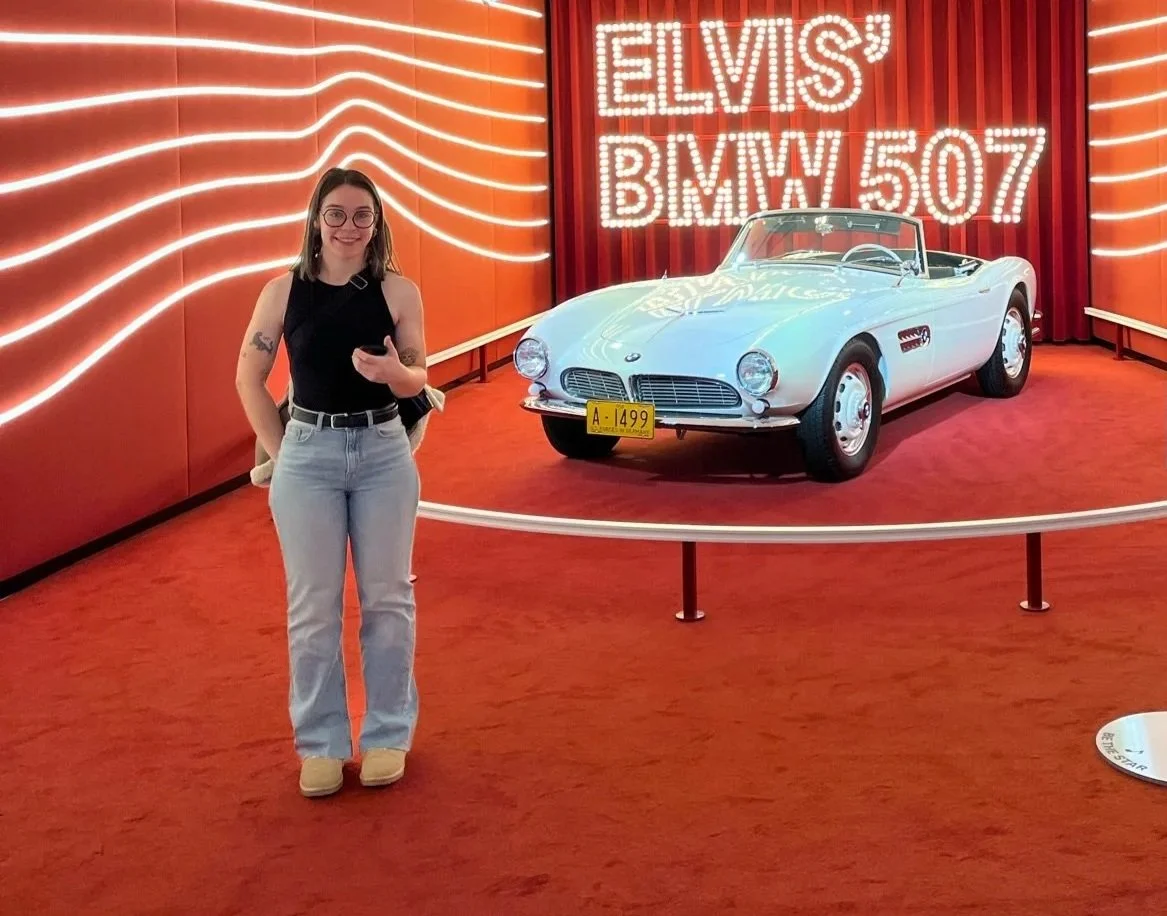Mujer con gafas y pantalones claros posando frente a un auto deportivo clásico blanco, en un exhibidor con fondo rojo y luces que forman letras que dicen 'ELVIS BMW 507'.