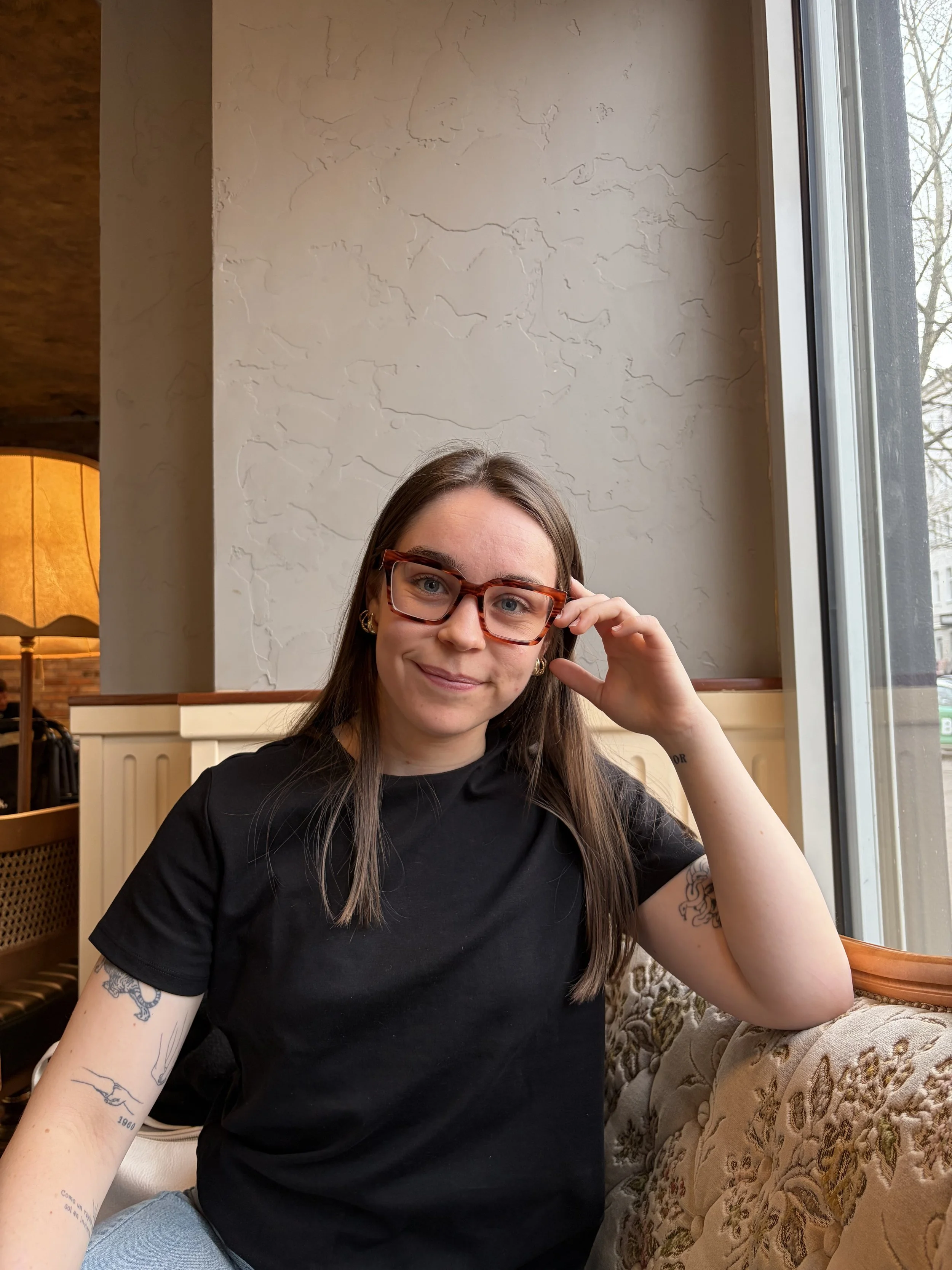 Mujer con gafas y pantalones claros posando frente a un auto deportivo clásico blanco, en un exhibidor con fondo rojo y luces que forman letras que dicen 'ELVIS BMW 507'.
