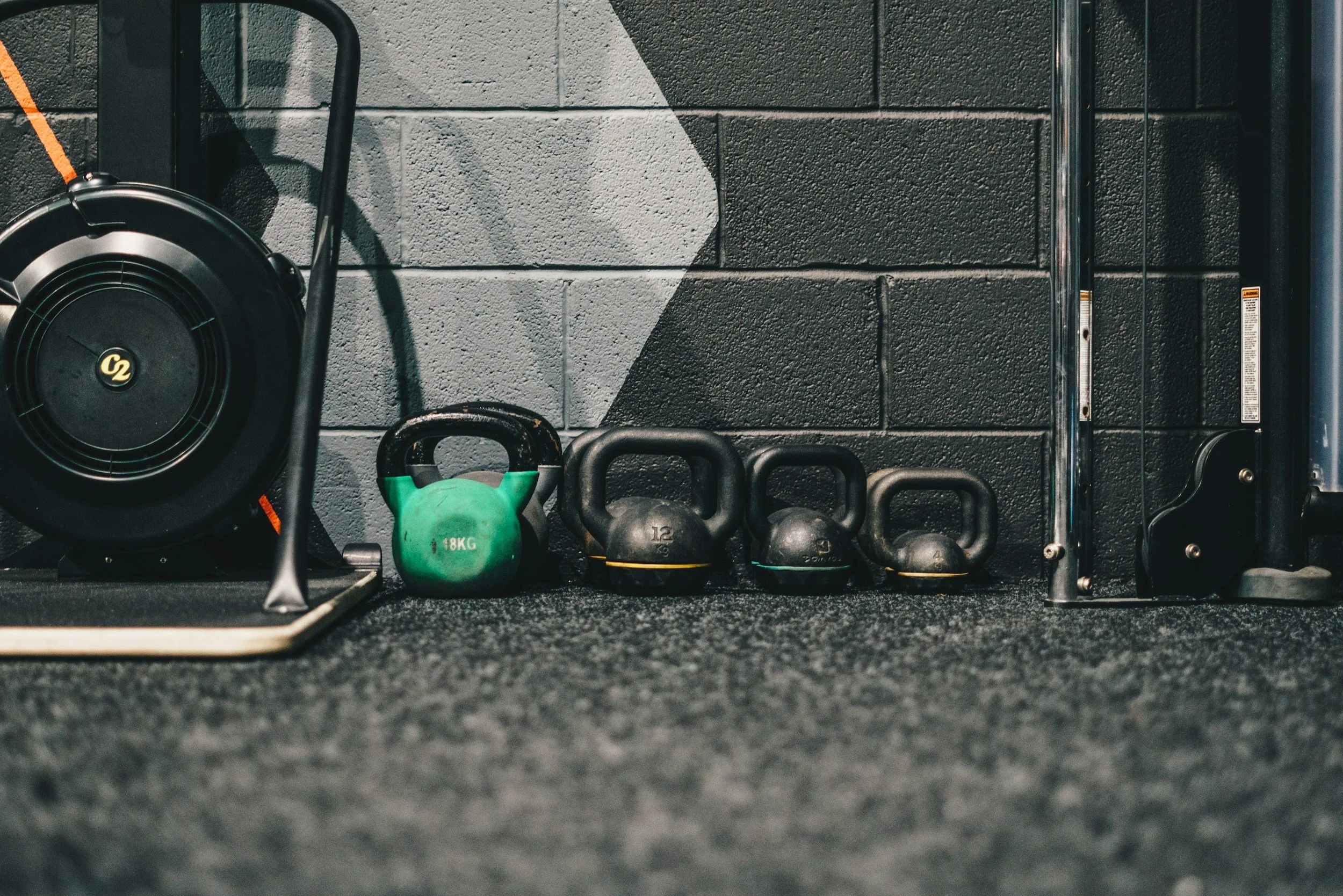 Gym wall with black and gray bricks and workout equipment including a SkiErg and a set of kettlebells on a gym floor.