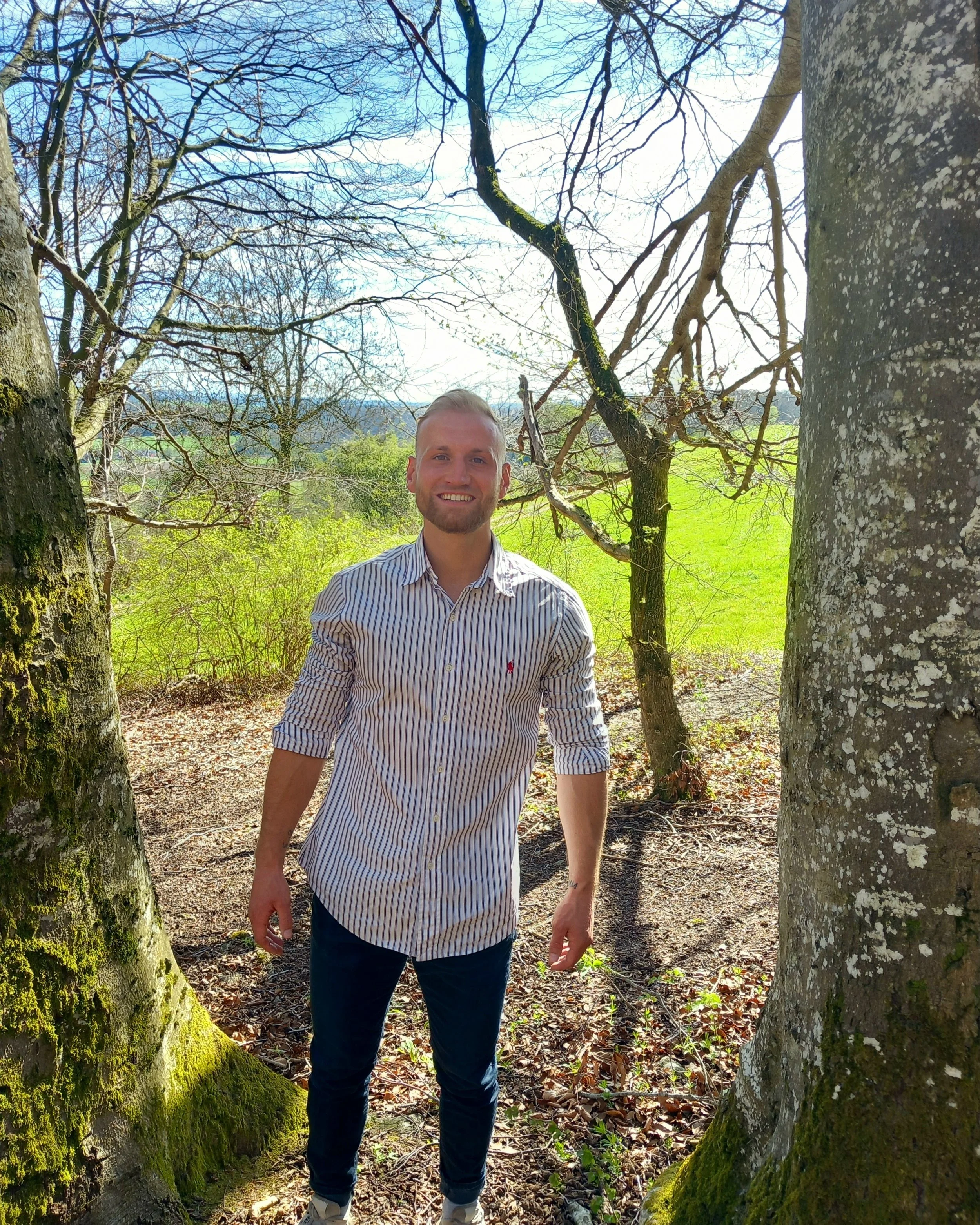 Ein lachender Mann steht in einem Wald zwischen Bäumen mit grüner Wiese im Hintergrund, bei sonnigem Wetter.