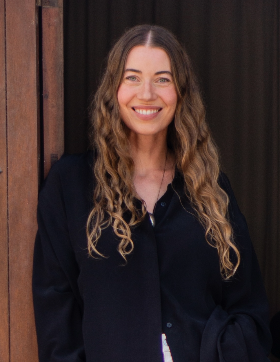 A woman with long, wavy brown hair wearing a black shirt, smiling and standing in front of a wooden door.