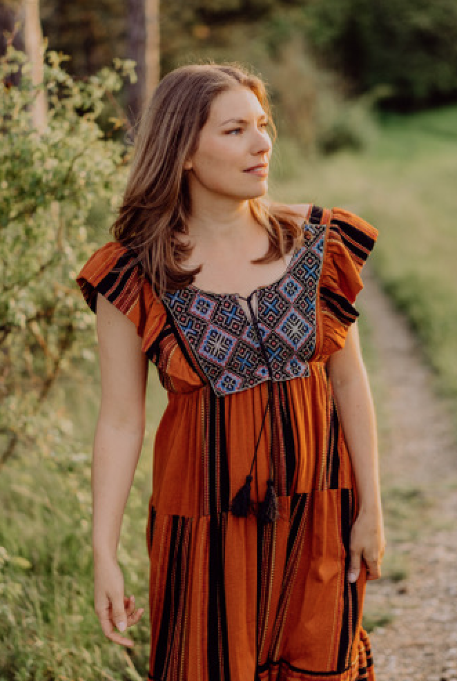 A woman with brown hair wearing an orange dress with blue and black embroidered details walking outdoors on a dirt path surrounded by green grass and trees.