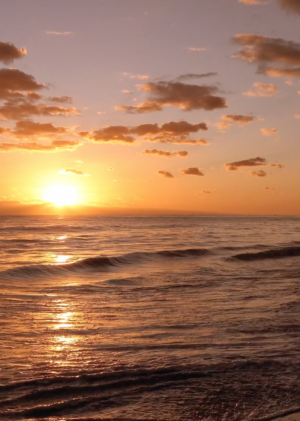 Sunset over the ocean with scattered clouds and gentle waves.