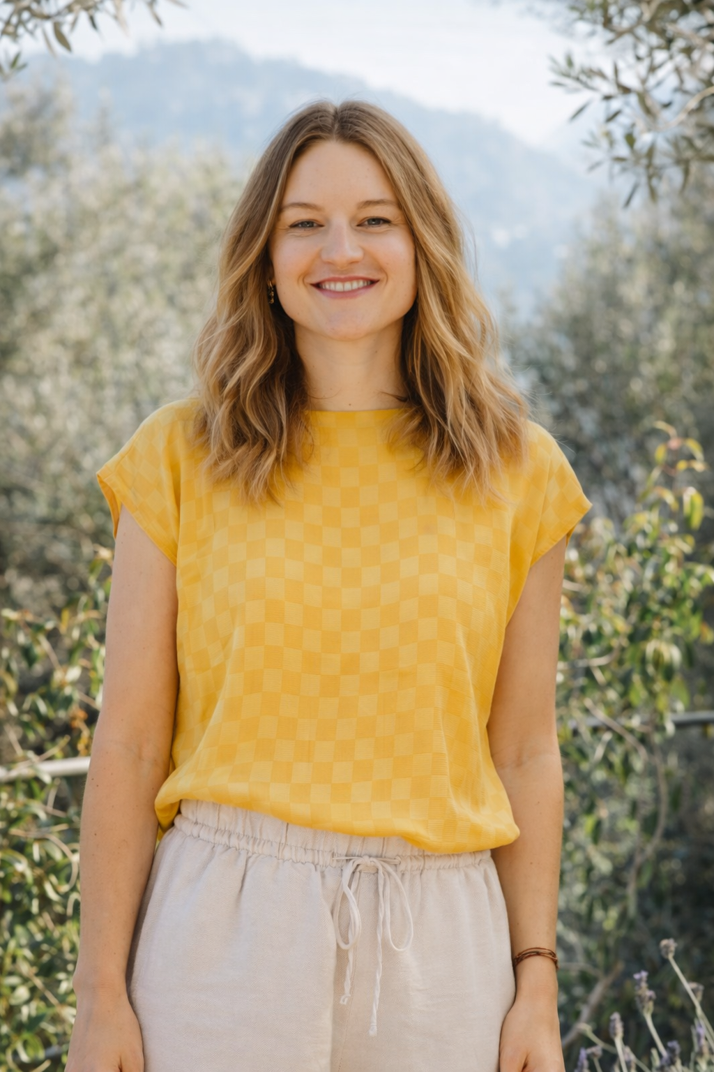 Smiling young woman with wavy golden blond hair wearing a yellow silk top and beige pants outdoors with mountains in the background.
