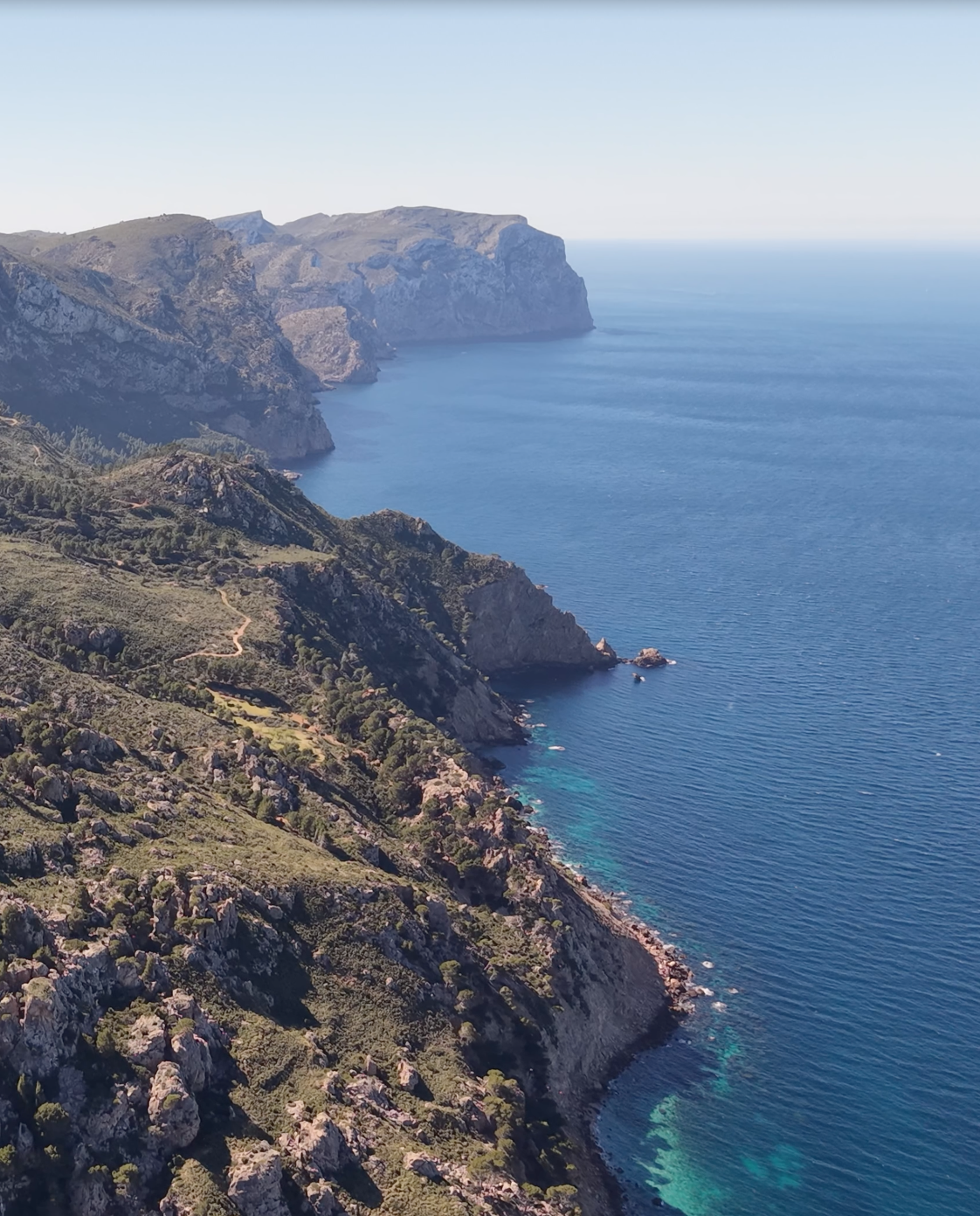 Aerial view of rugged Tramuntana coastline with cliffs, green vegetation, and blue ocean water extending to the horizon.