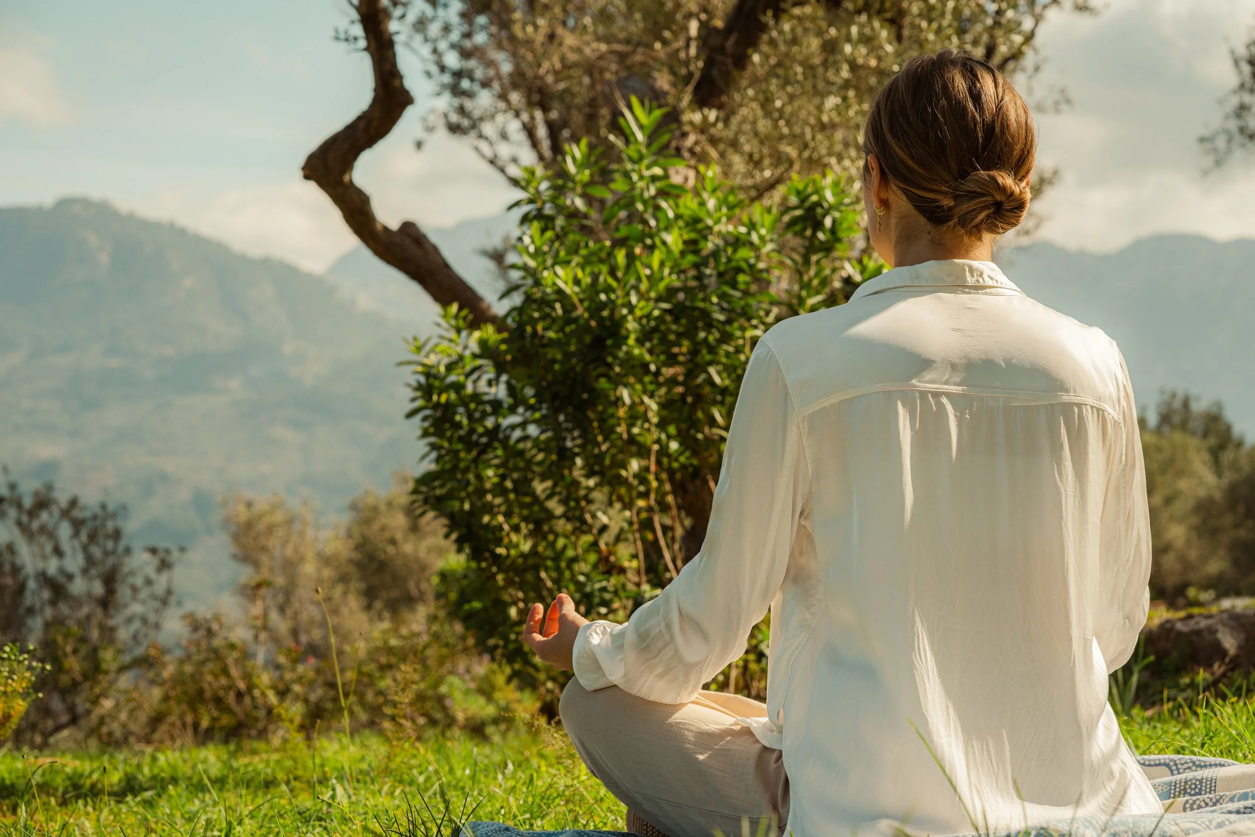 A woman in a white shirt practicing meditation outdoors on a grassy area with mountains and trees in the background.