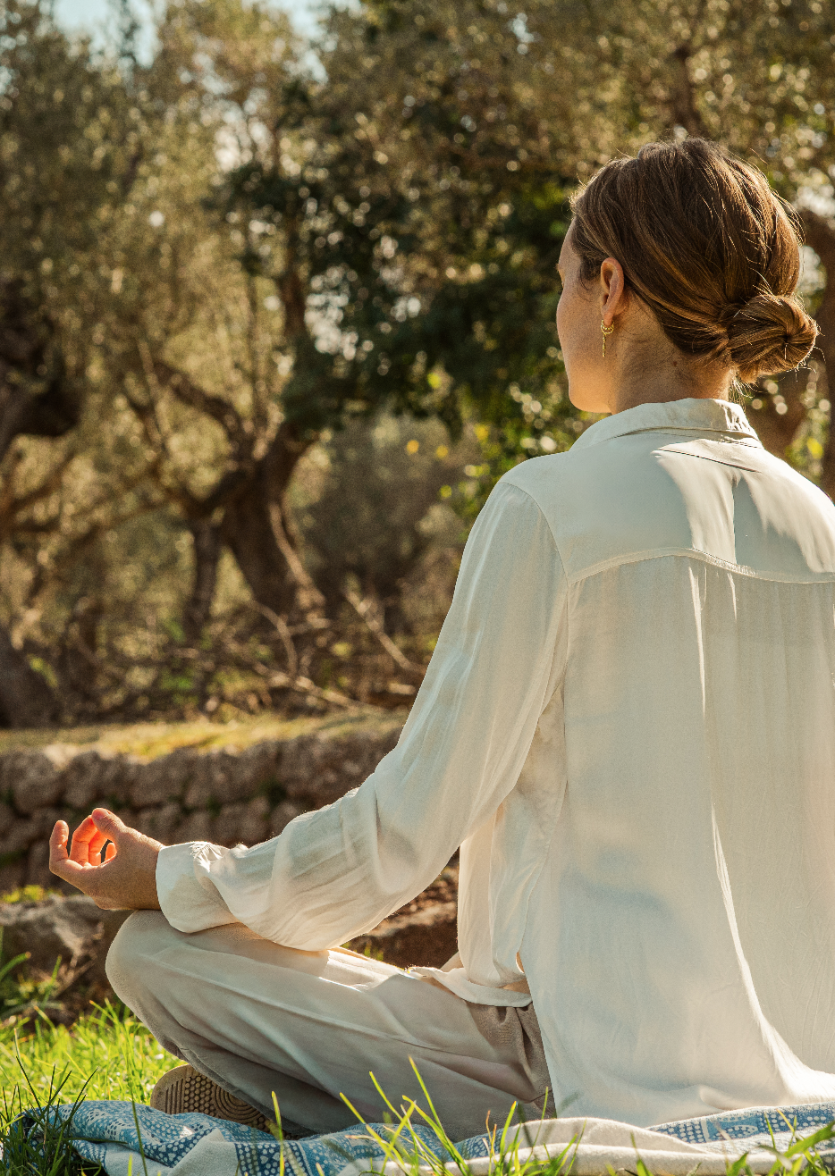 A woman practicing yoga outdoors, sitting in a cross-legged position on a blanket, with her hand in a mudra, in a natural setting with trees and sunlight.
