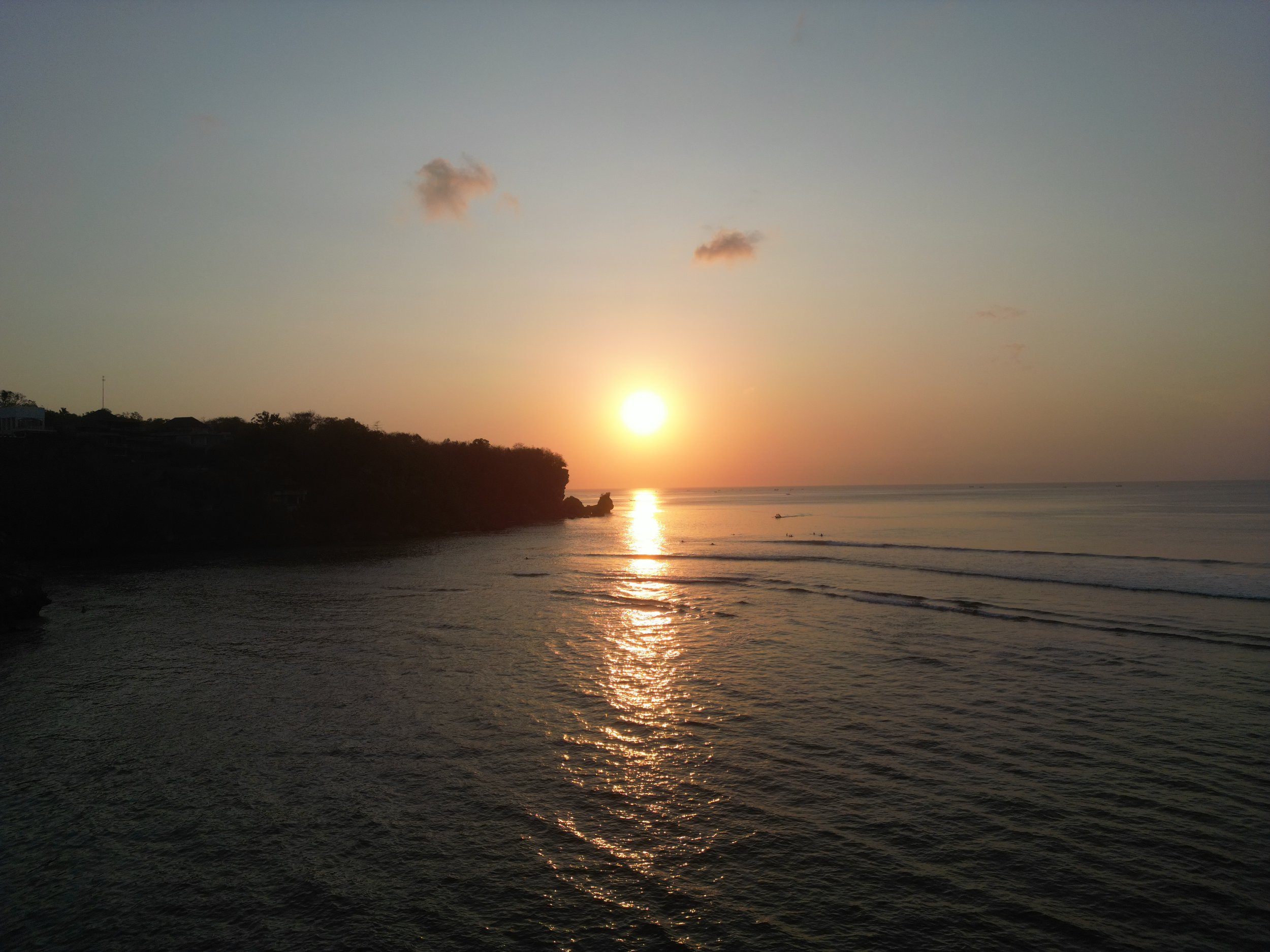 Sunset over the ocean with silhouettes of trees and some small boats in the water.