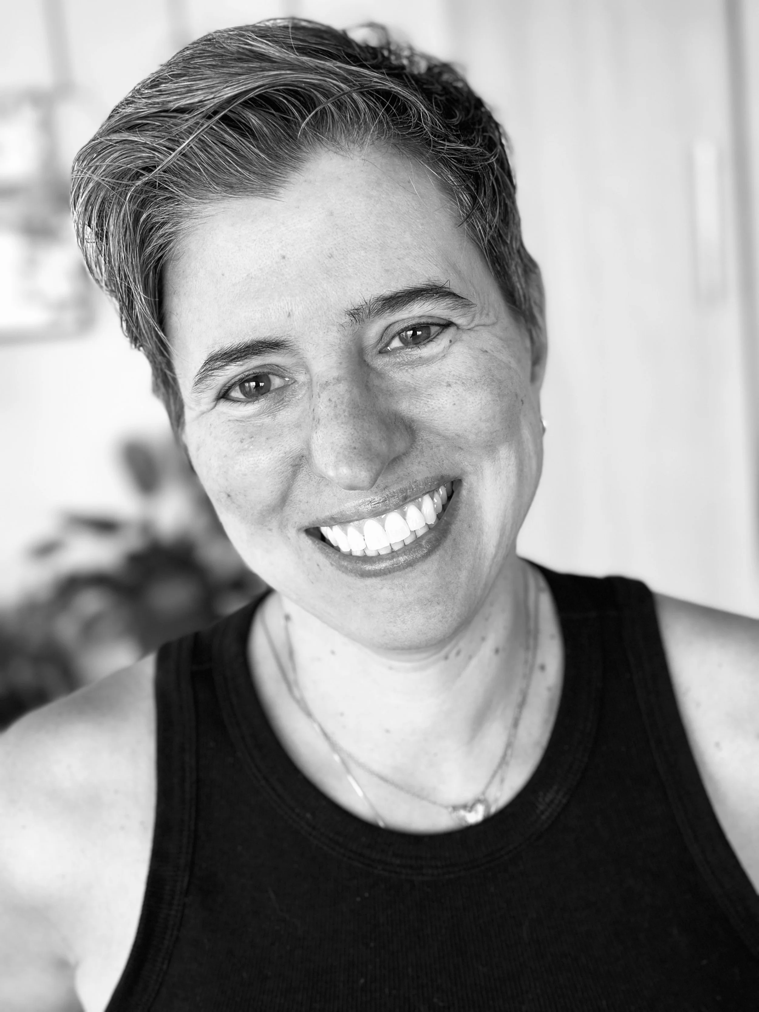 Black and white close-up photo of a smiling woman with short, styled hair, wearing a black tank top and a necklace.