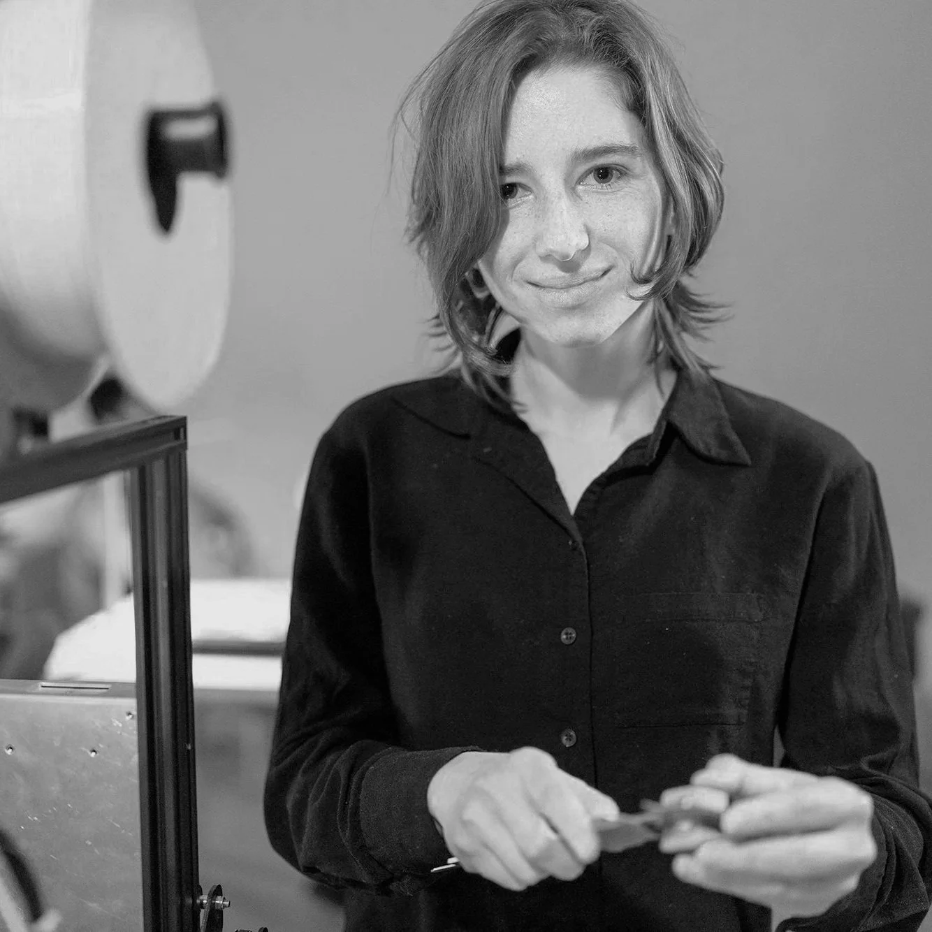 A woman with medium-length hair, wearing a dark shirt, is looking at a flat surface, possibly a mirror or a screen, with a slight smile. She is indoors with a plain background and appears to be focusing on something in front of her.