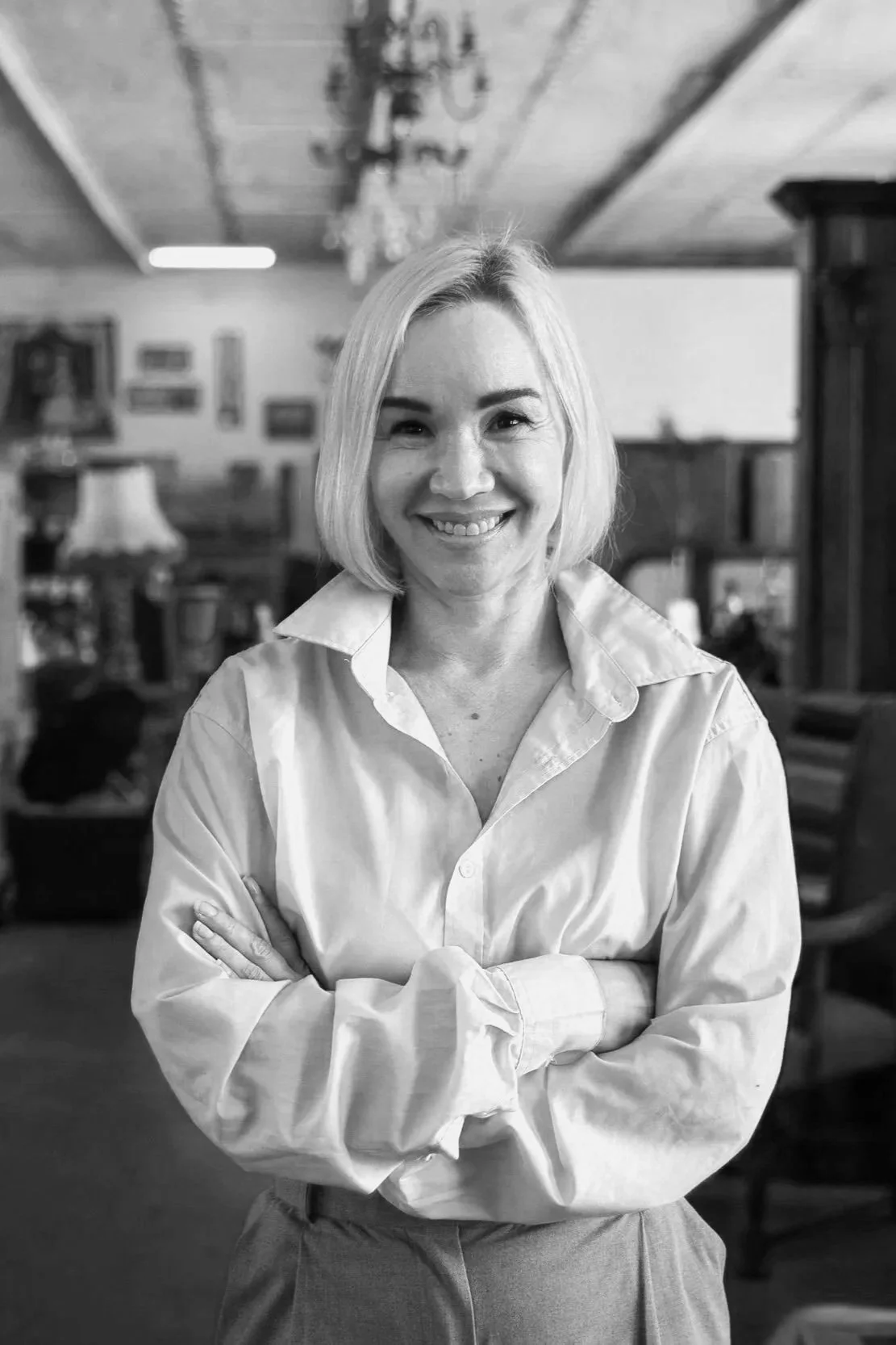 Black and white photo of a woman with blonde hair wearing a button-up shirt, smiling with arms crossed, indoors with furniture and pictures on the wall in the background.