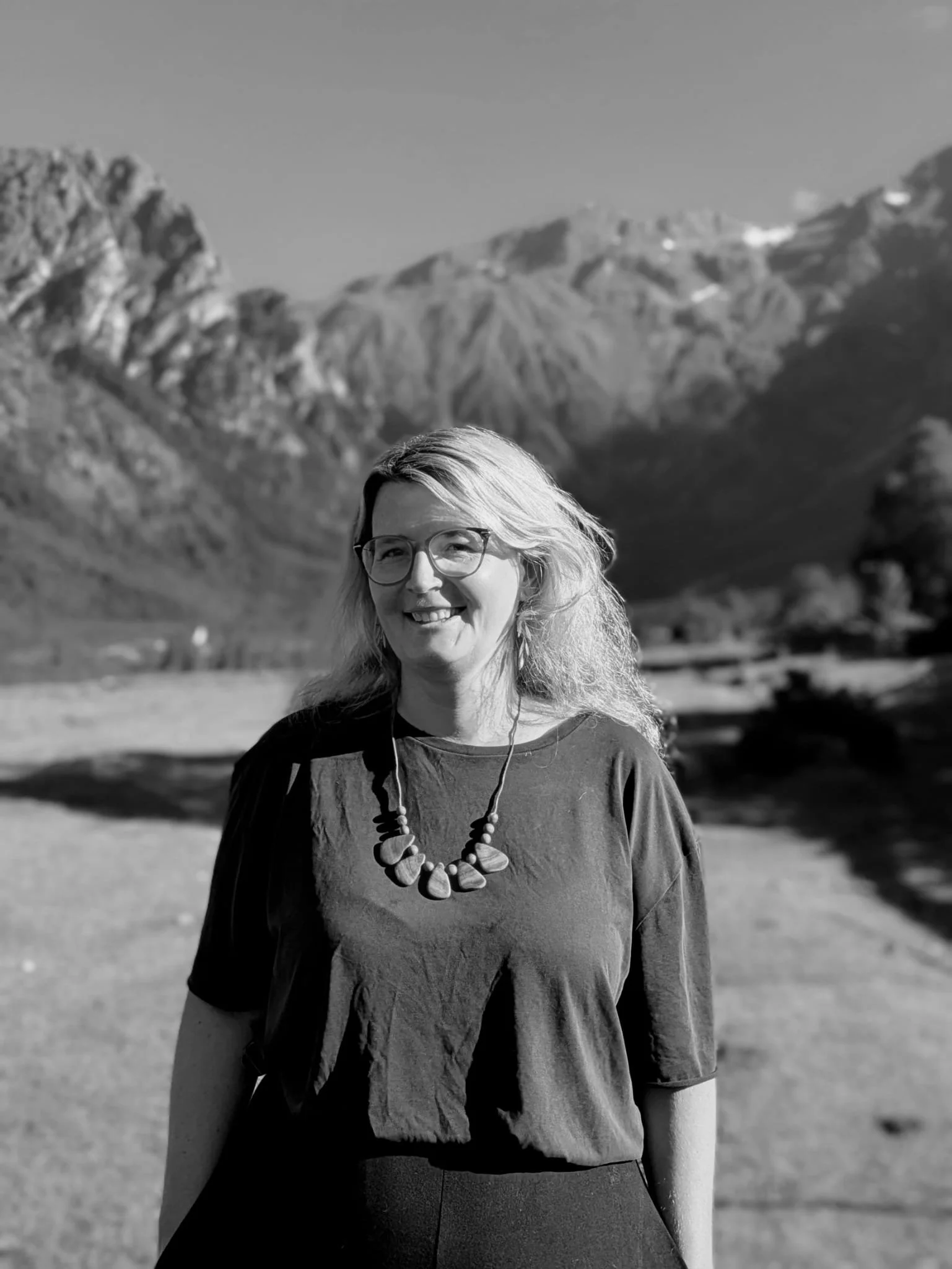 Black and white photograph of a smiling woman with glasses, wearing a necklace and a short-sleeved blouse, standing outdoors with mountains in the background.