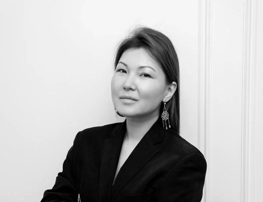 A black and white portrait of a woman with straight hair wearing earrings and a black blazer, standing against a plain background.