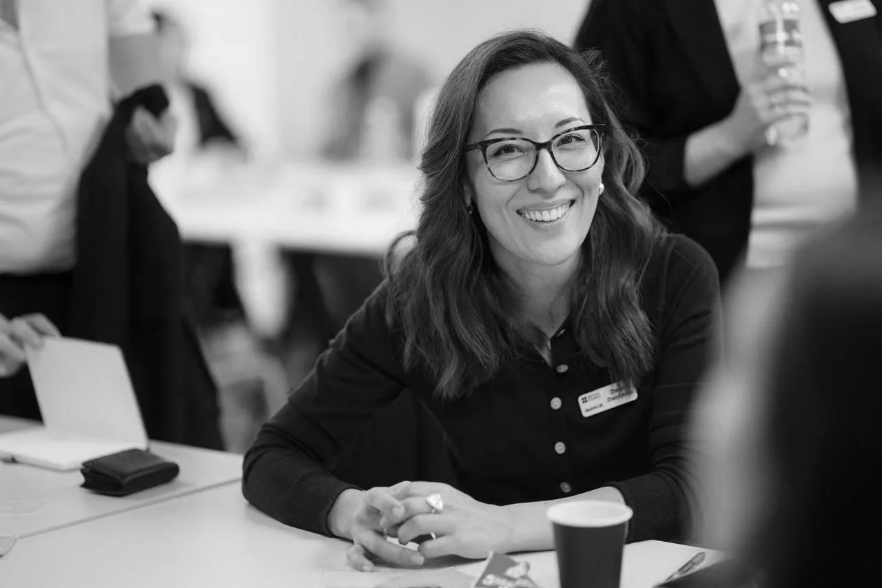 A woman with glasses smiling sitting at a table during a social or professional event, with people and tables in the background.