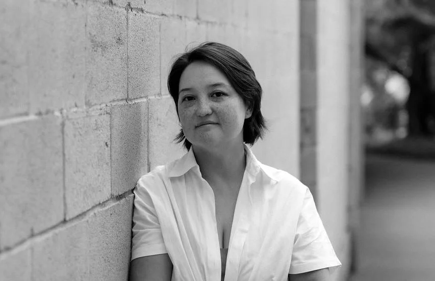A woman with short dark hair, wearing a white shirt, leaning against a brick wall on an outdoor path in black and white.