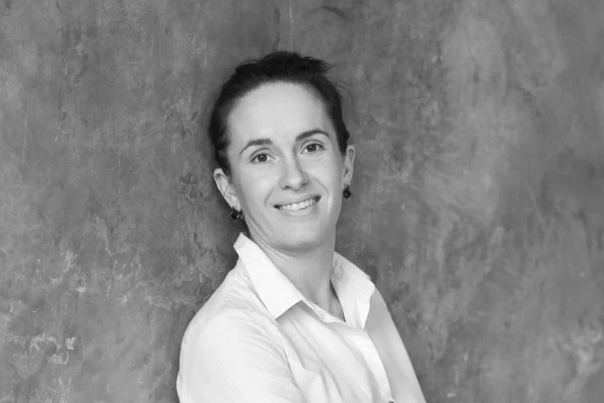 Black-and-white photo of a woman with dark hair, smiling, wearing a collared shirt, standing against a textured wall.