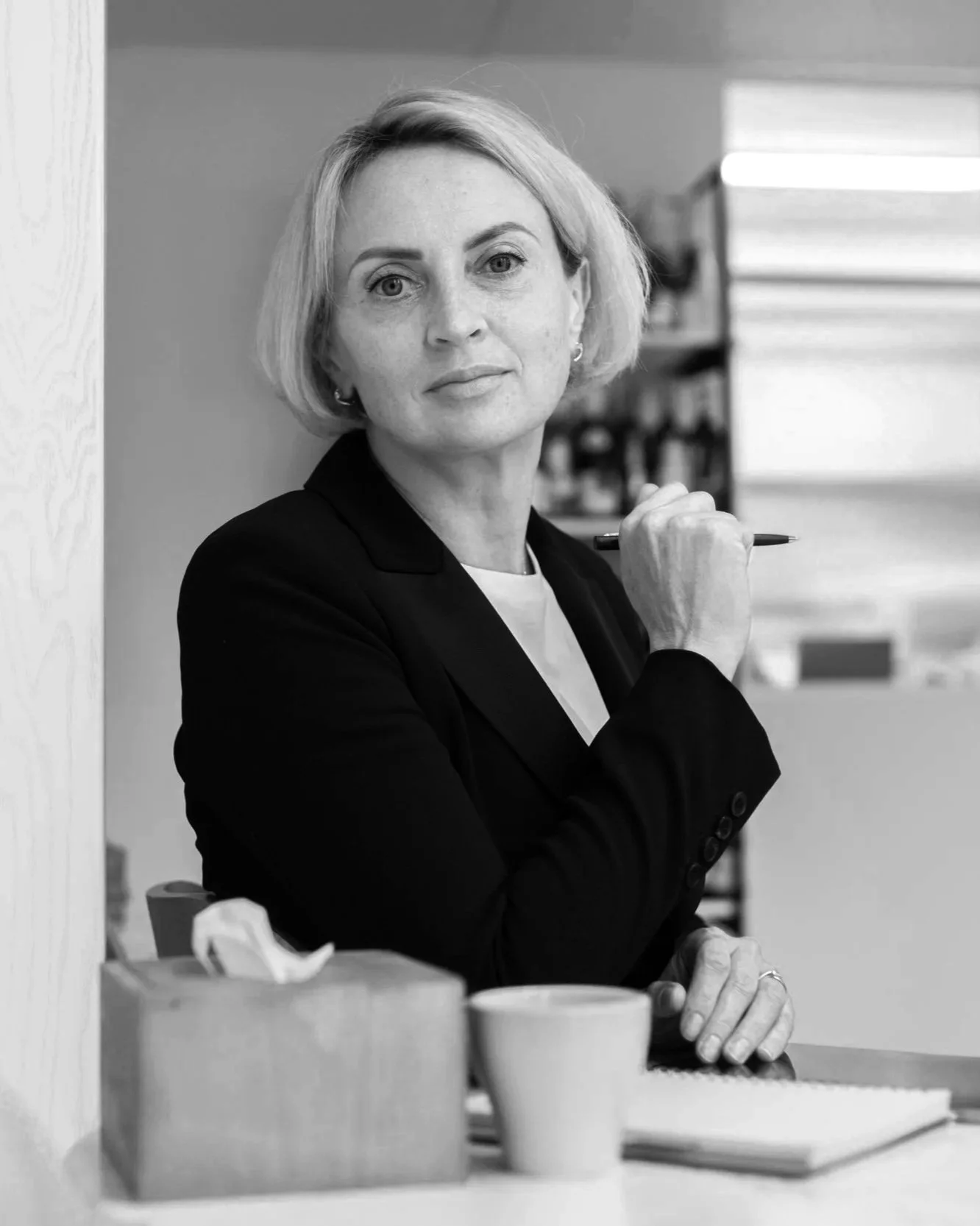 Black and white photo of a woman with short blonde hair, wearing a dark blazer, sitting at a table, holding a pen near her chin, with a bookshelf in the background and coffee cups on the table.