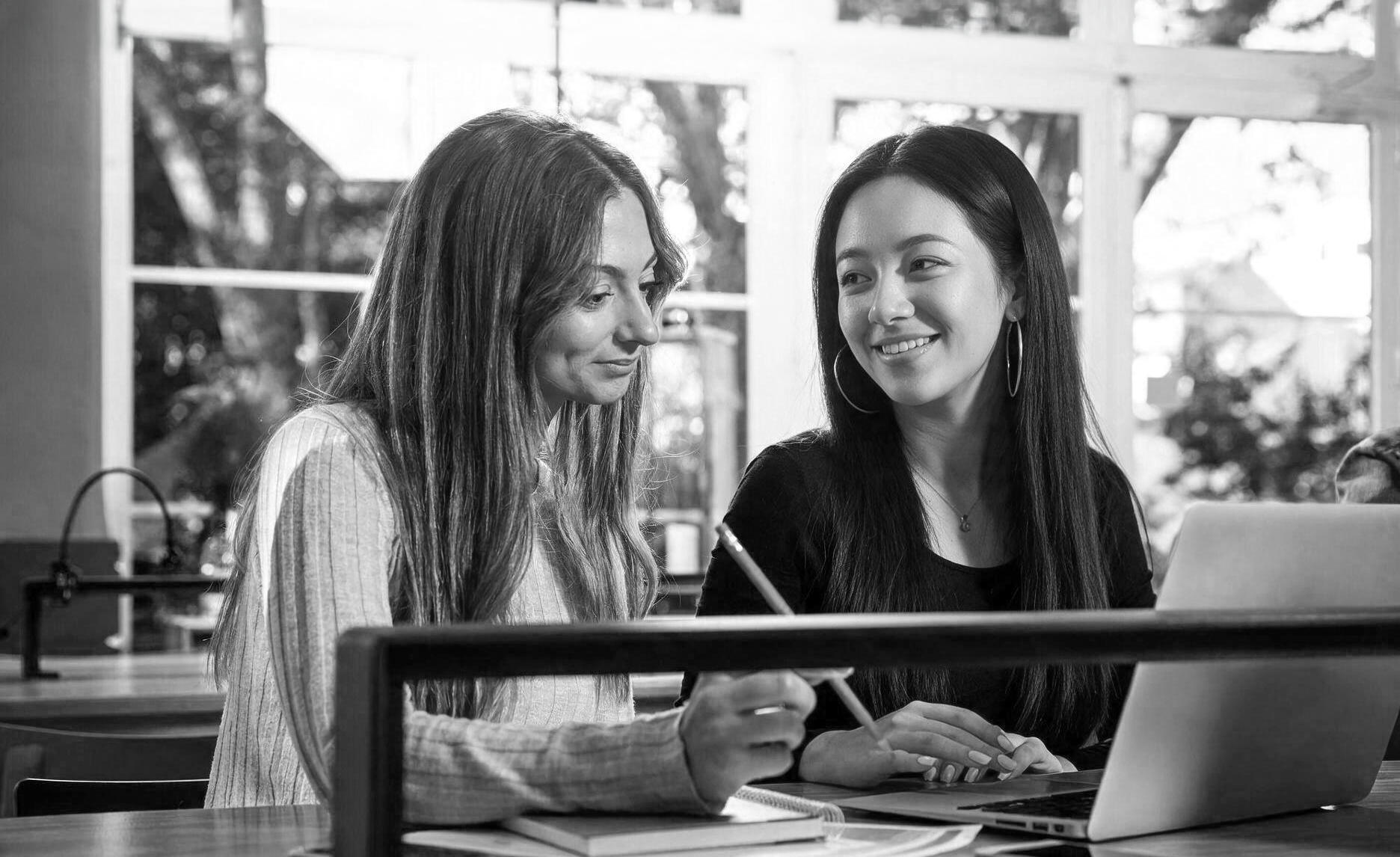 Two women sitting at a desk in a bright room with large windows, smiling and looking at a laptop.