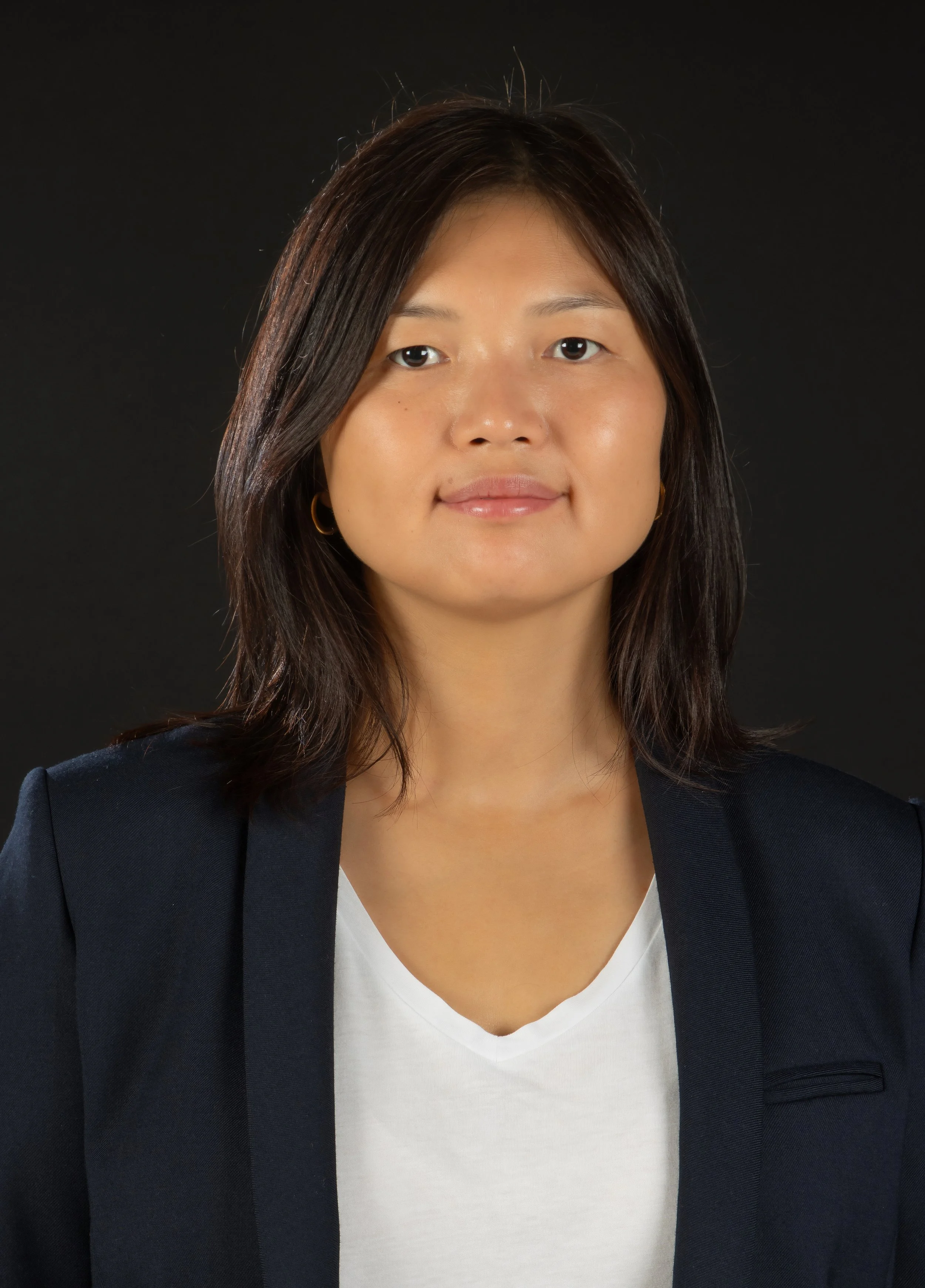 Portrait of a woman with dark brown hair, wearing a navy blazer and white top, against a black background.
