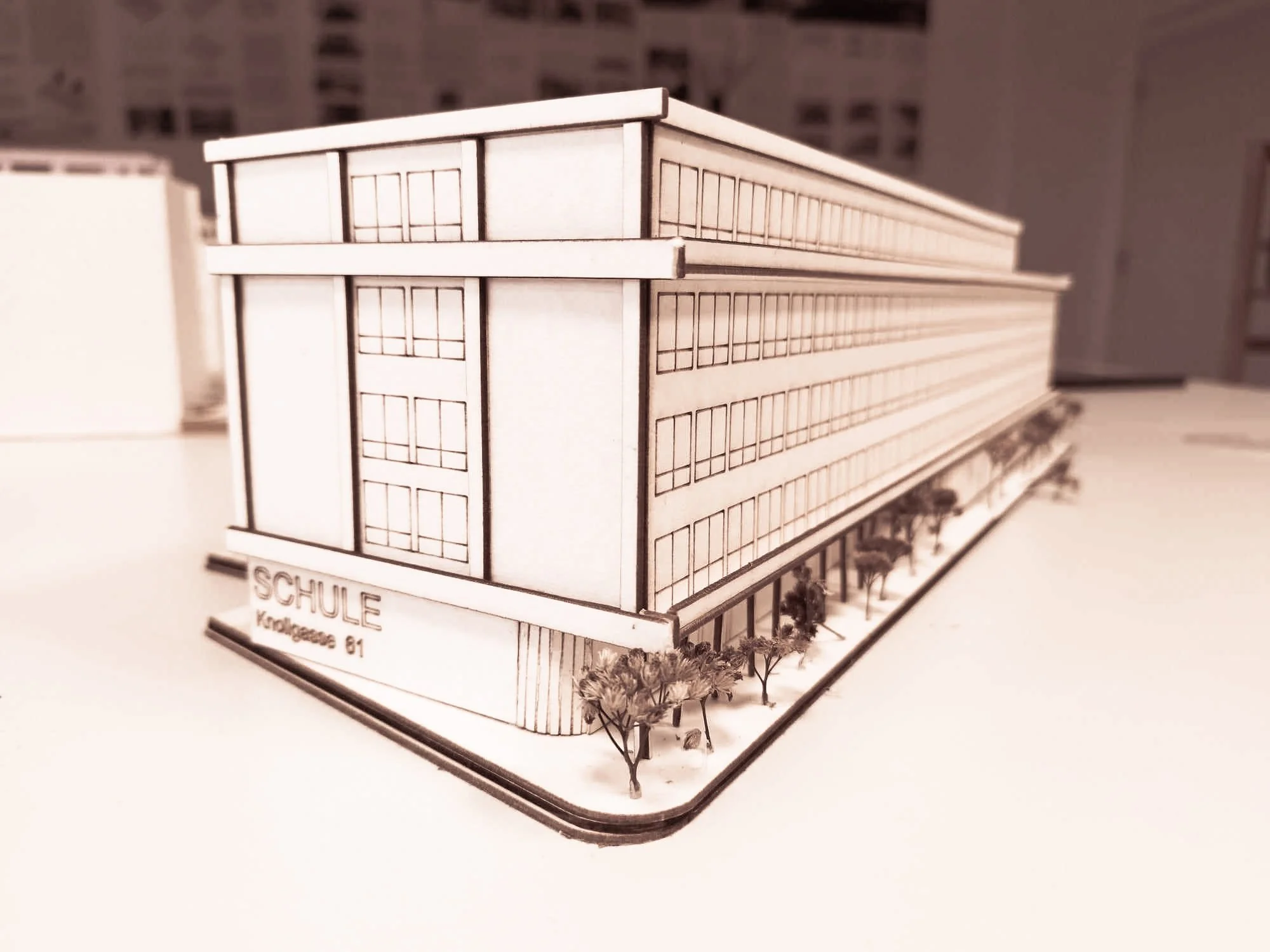 Architectural model of a school building with multiple floors, labeled 'SCHULE' and 'Klingeasie 81', with small trees around the base.