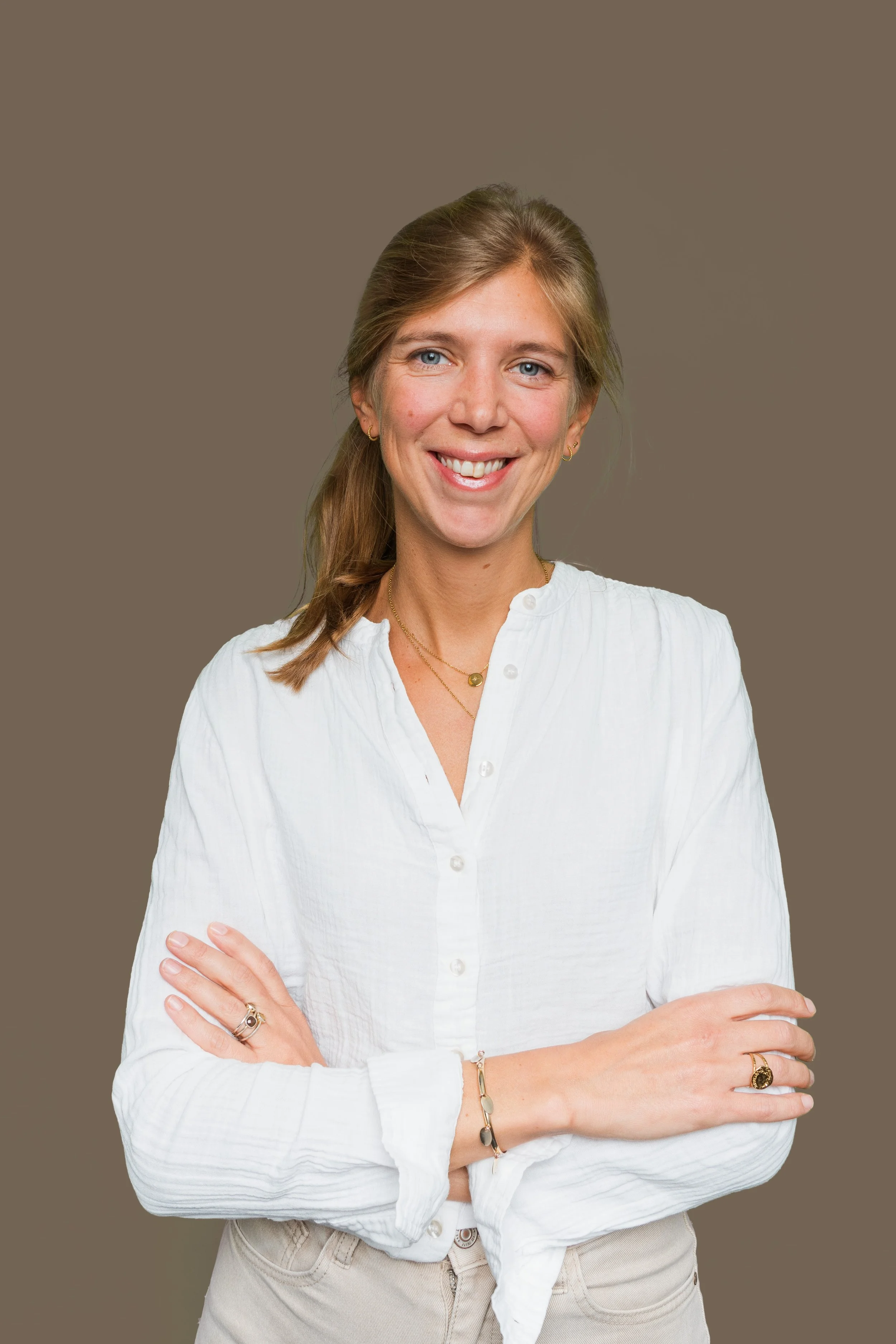 Une femme souriante portant une chemise blanche et des bijoux, posant les bras croisés devant un fond uni beige.