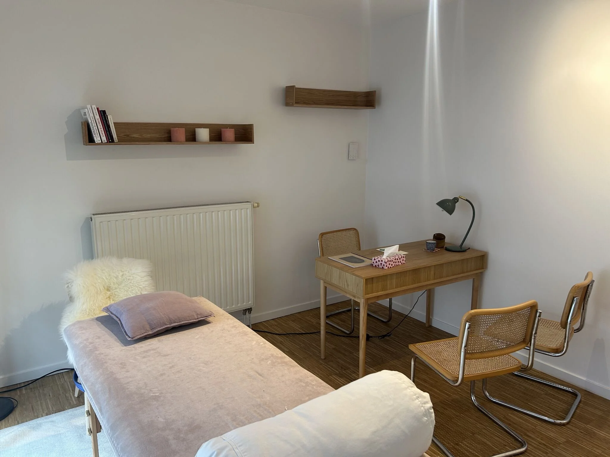 Une pièce avec une table de soin, une table en bois avec une lampe, et deux chaises en bois avec assise en osier. Deux étagères murales en bois, une avec des livres et des bougies, l'autre vide. Le décor est minimaliste avec des tons neutres.