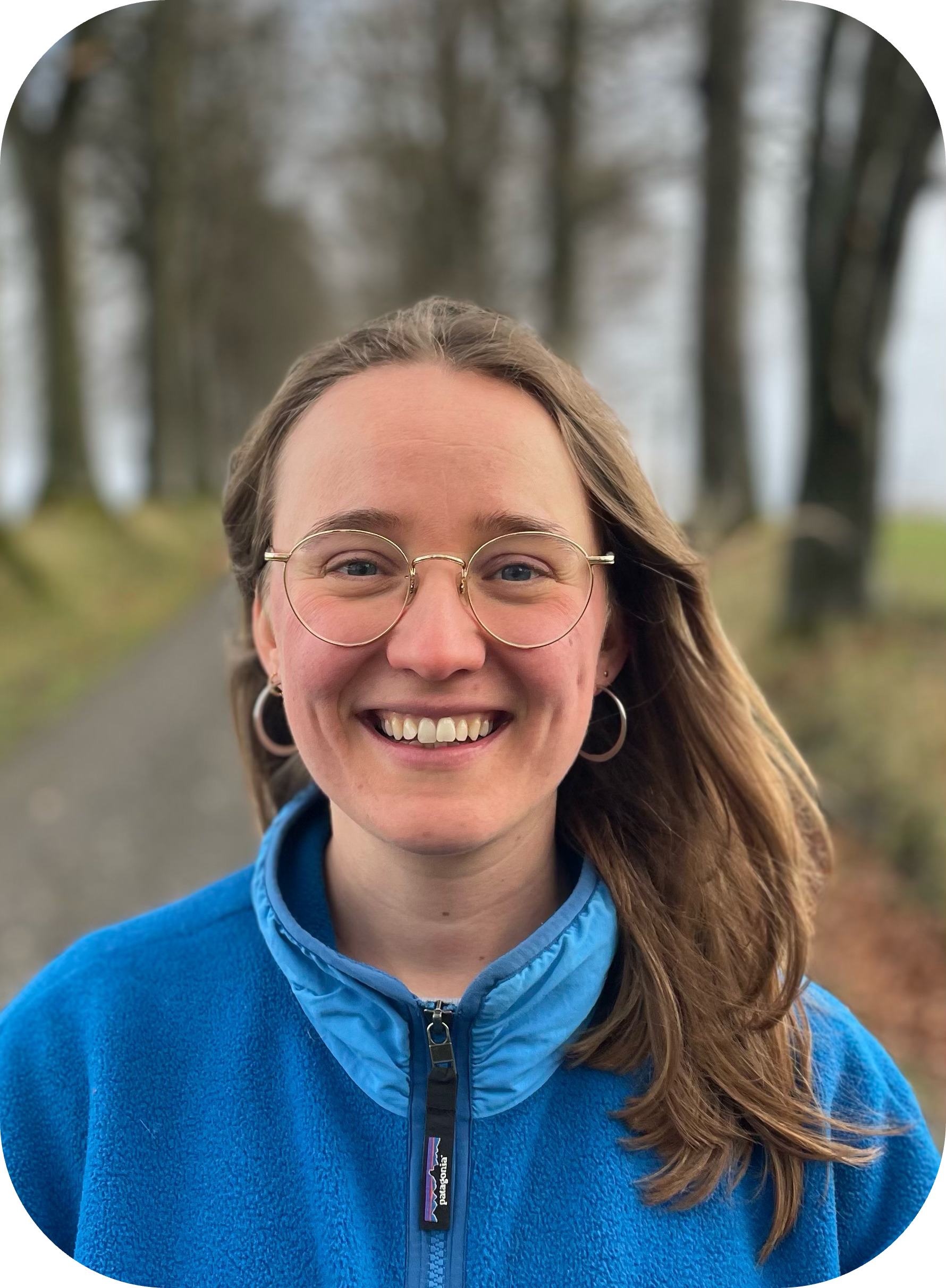 Jeune femme souriante portant des lunettes rondes et un pull bleu, debout sur un chemin forestier.