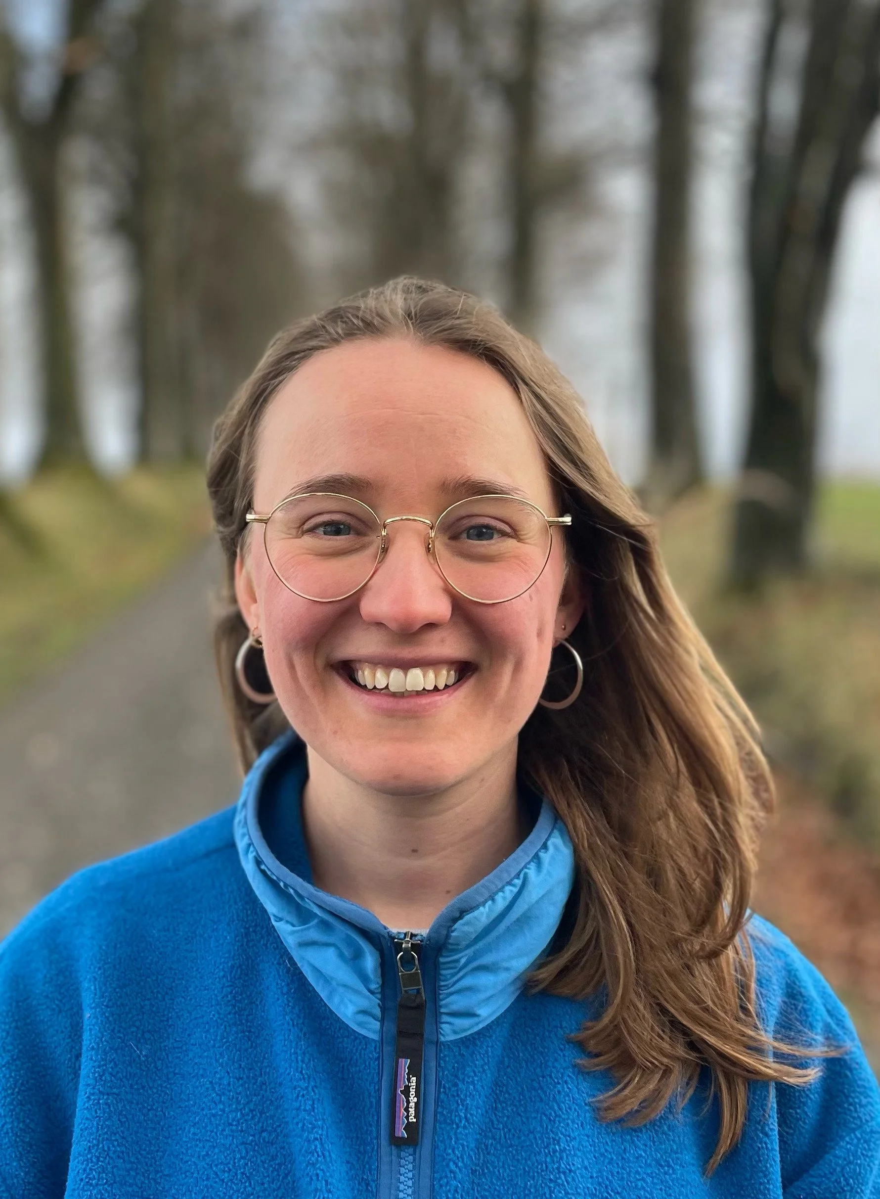 Une femme souriante portant des lunettes rondes et un pull bleu, dans un environnement extérieur avec des arbres et un chemin derrière elle.
