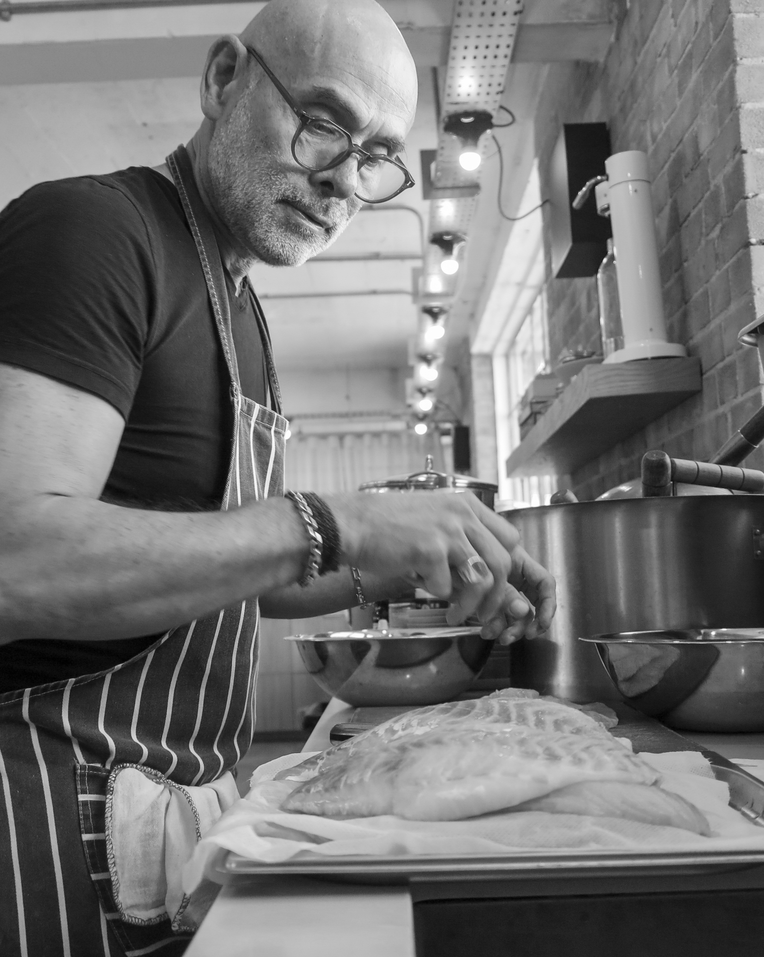 A man with glasses and a shaved head wearing a striped apron preparing food in a kitchen. He is working on a large piece of dough on a cloth-covered surface, with kitchen utensils and a food processor nearby. The kitchen has a brick wall and wooden shelves.