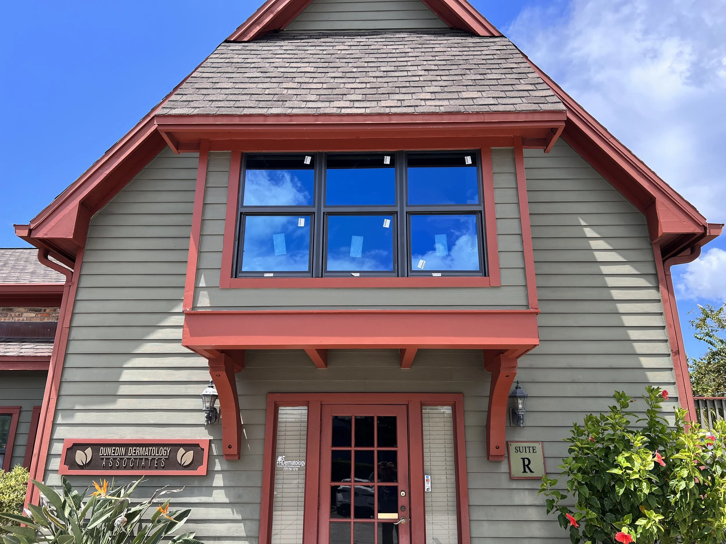 The front of a building with green siding and red trim, with a large window above the entrance reflecting the blue sky and clouds, and a sign that reads 'Dunedin Dermatology Associates.' There are plants and flowers in front of the building.